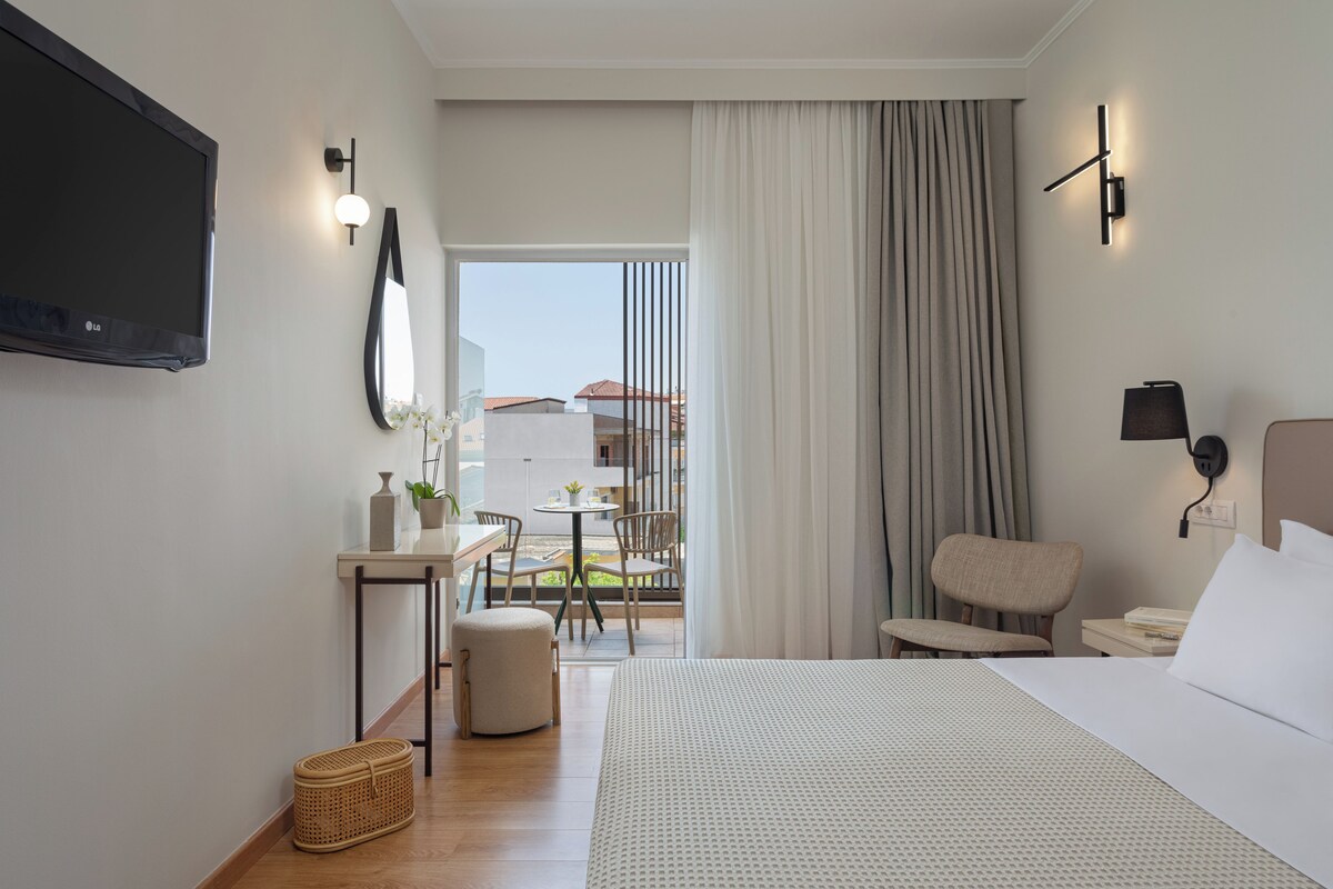 A hotel room with a bed, a desk with a chair, a TV mounted on the wall, and a view of the outside balcony.