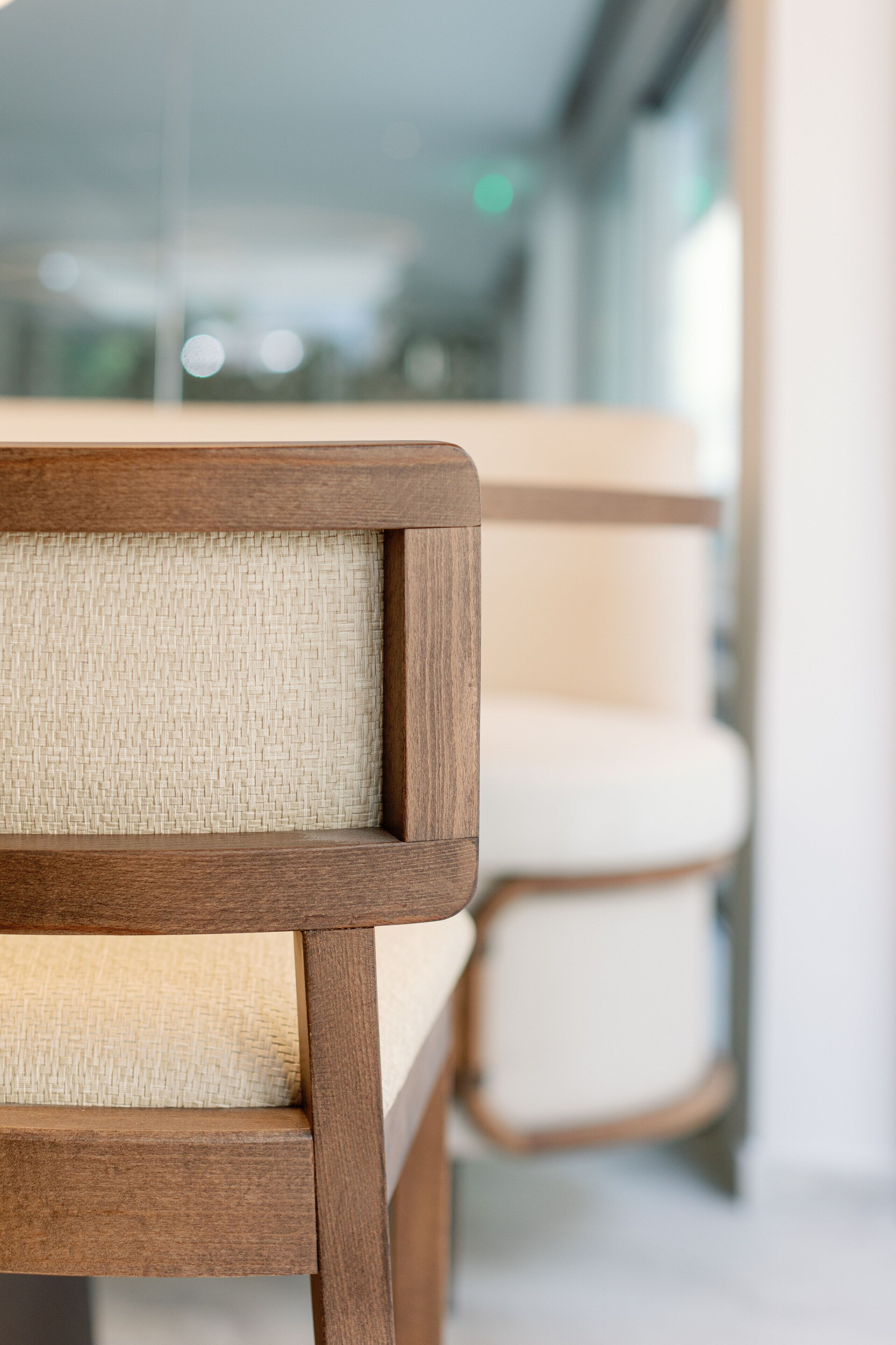Primer plano de una silla de madera con asiento y respaldo trenzados, parte de un interior moderno con separadores de vidrio y una paleta de colores neutros.