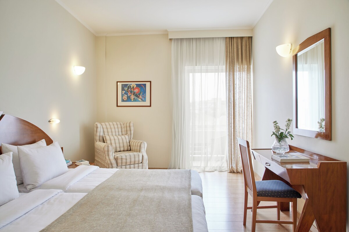 A hotel room with a large bed, a desk with a chair, a small table with a vase, and a painting on the wall.