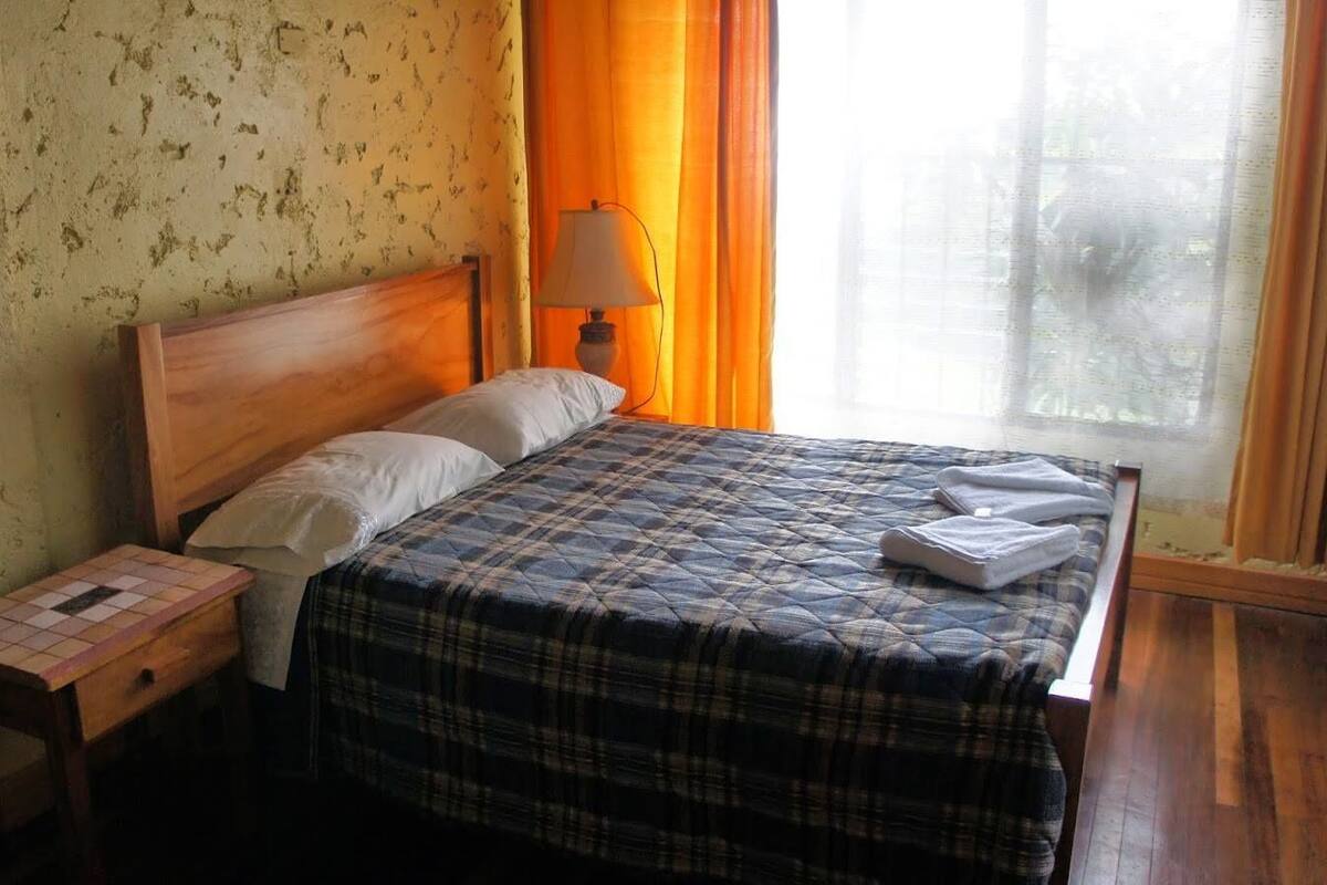 A single bed with a plaid bedspread, a wooden nightstand, a lamp, and a window with curtains.