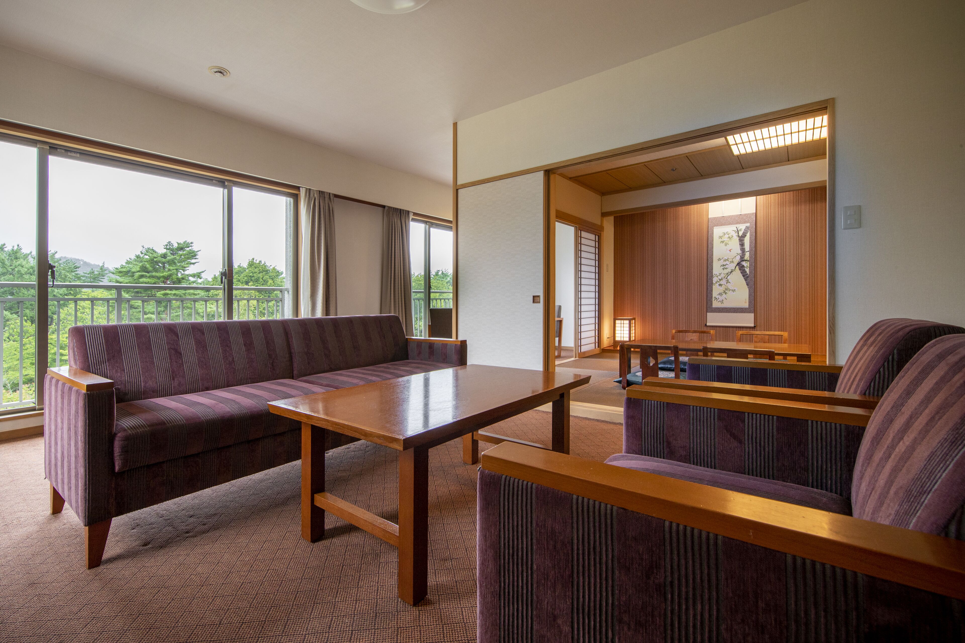 A modern hotel room with a large window, a sofa, a wooden table, and a view of the outdoors.
