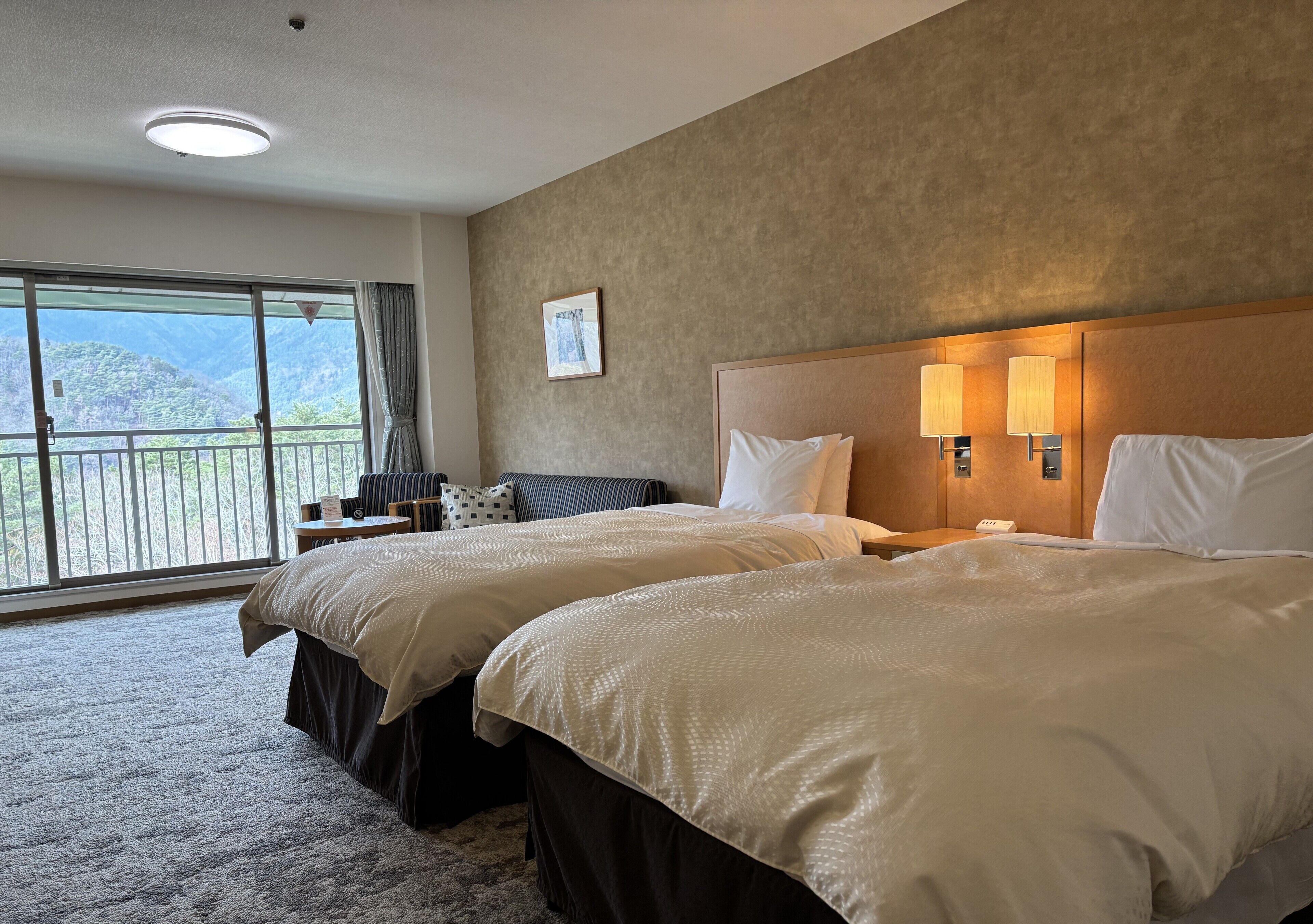 A hotel room with two beds, a large window with a view of mountains, and a balcony.