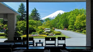 Une vue prise de l’intérieur d’un bâtiment, à travers une fenêtre donnant sur un jardin bien entretenu, avec une route, des arbres et une montagne en arrière-plan.