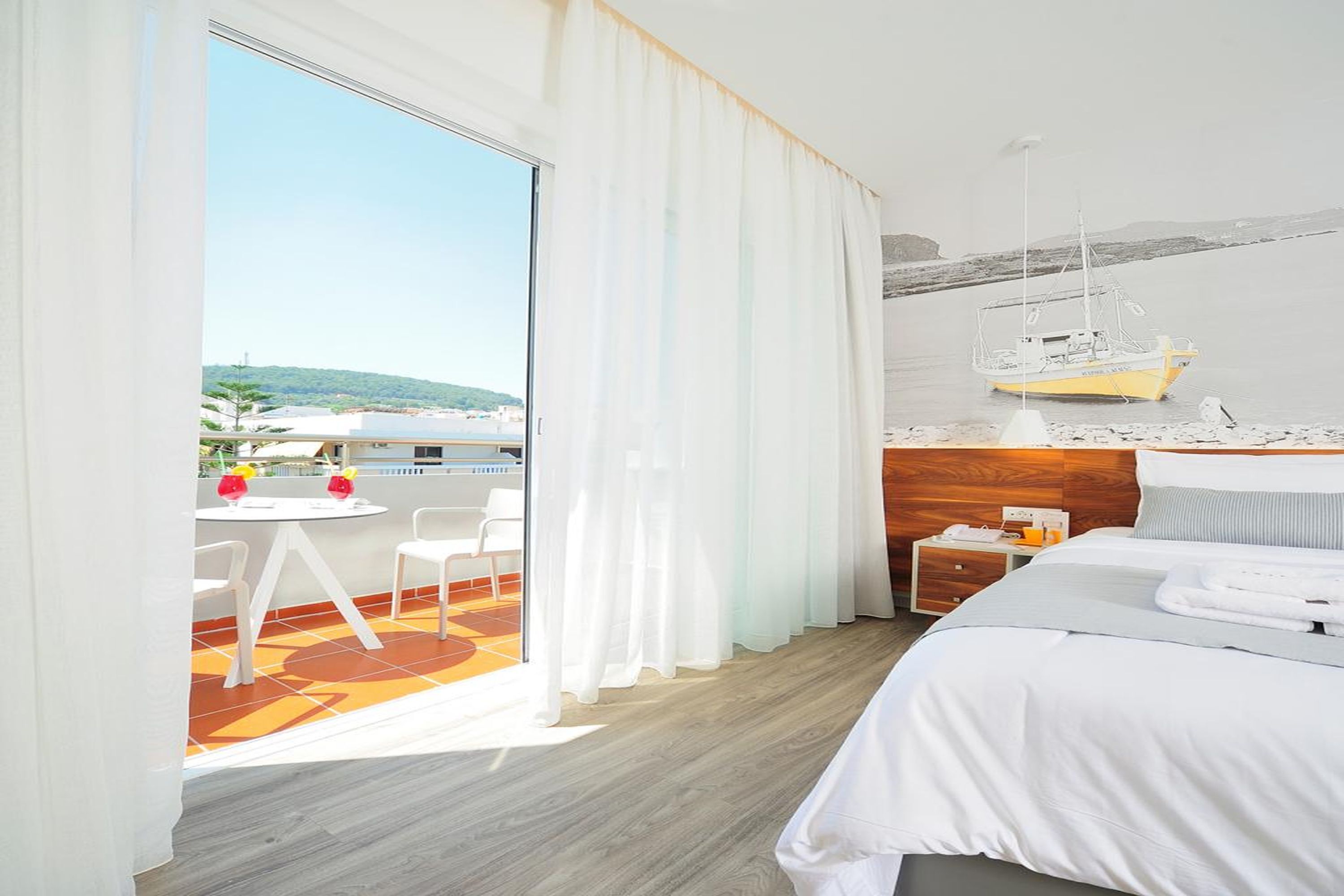 Camera d'albergo con un letto, un tavolino con sedie, un comodino e vista sul mare e su una barca.