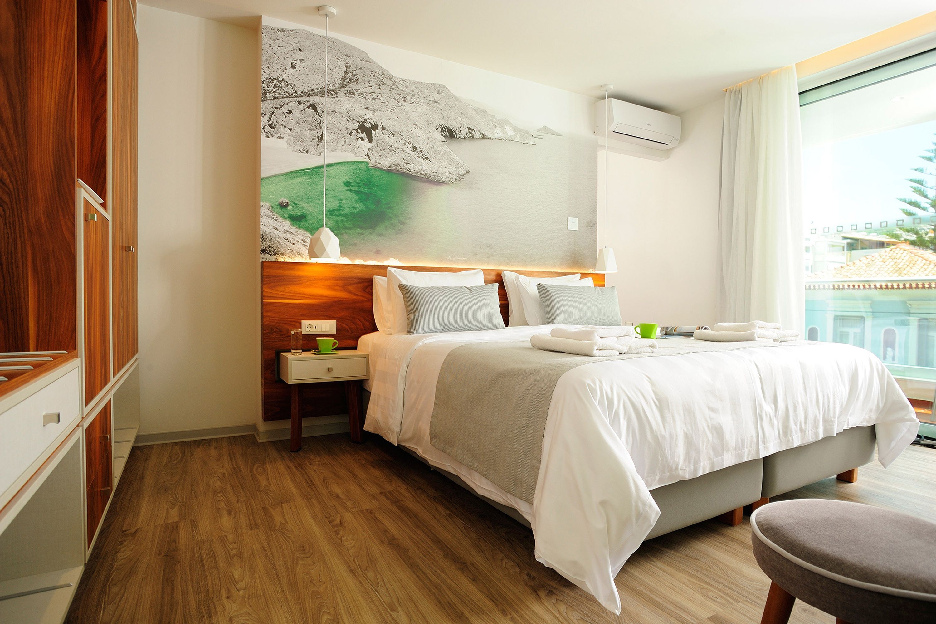 A hotel room with a bed, nightstands, a green cup, and a view of a building through the window.