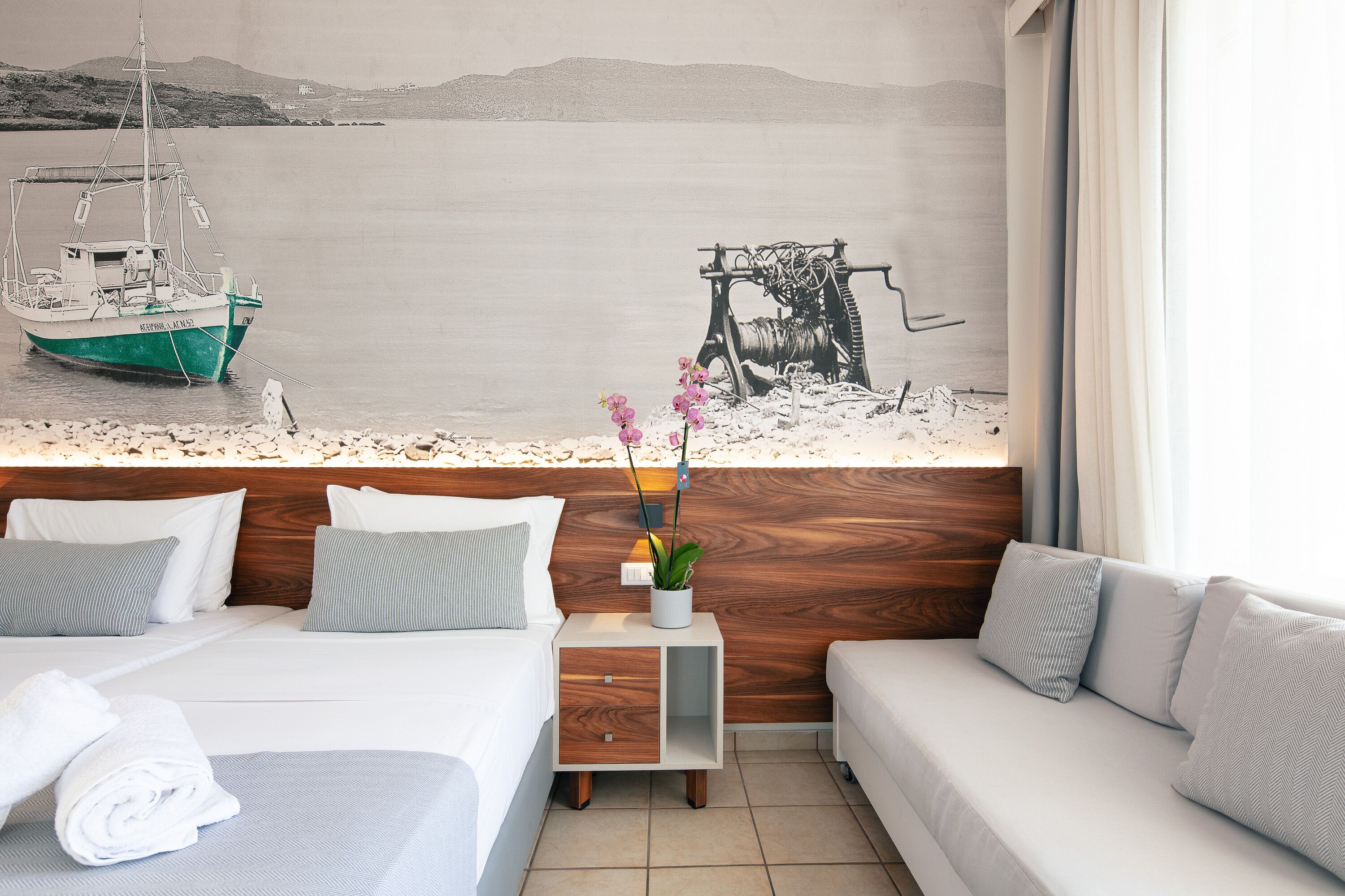 A hotel room with a bed, a sofa, a nightstand with a plant, and a mural of a boat and machinery on the wall.