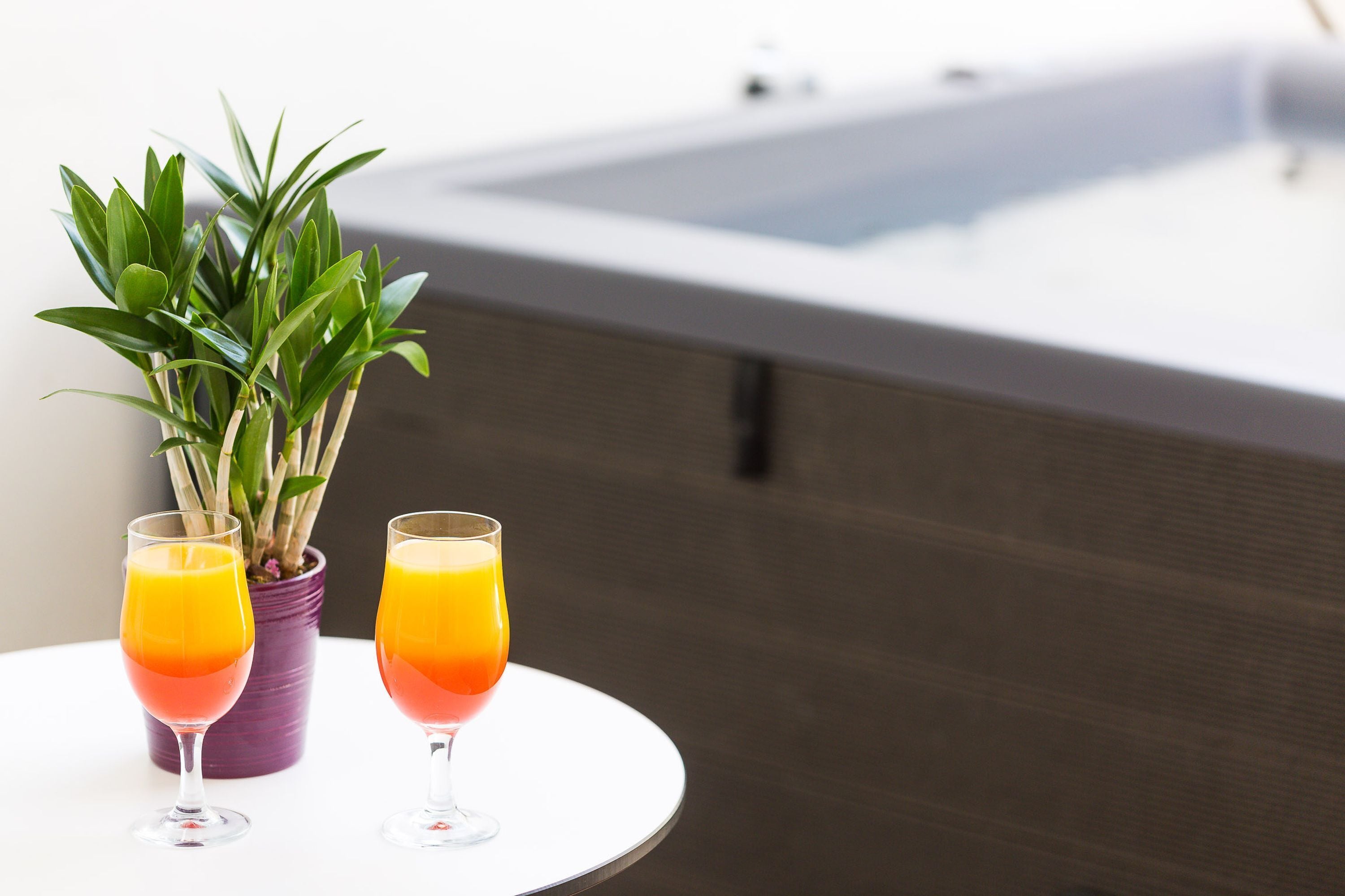 Deux verres de jus d’orange sont posés sur une table blanche, à côté d’une plante en pot.