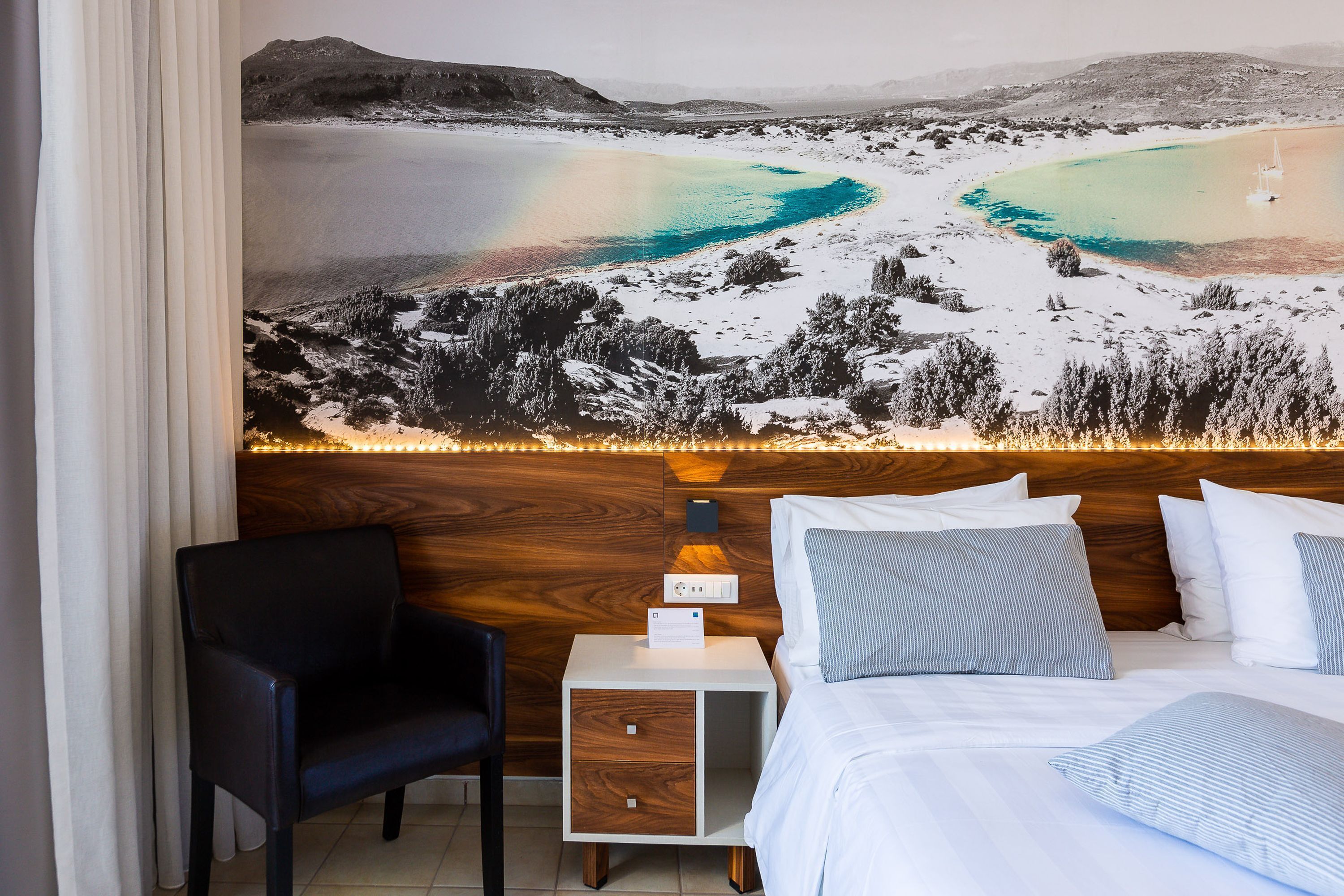 Ein Hotelzimmer mit einem Bett, einem Nachttisch, einem Sessel und einem großen Landschaftsbild an der Wand.