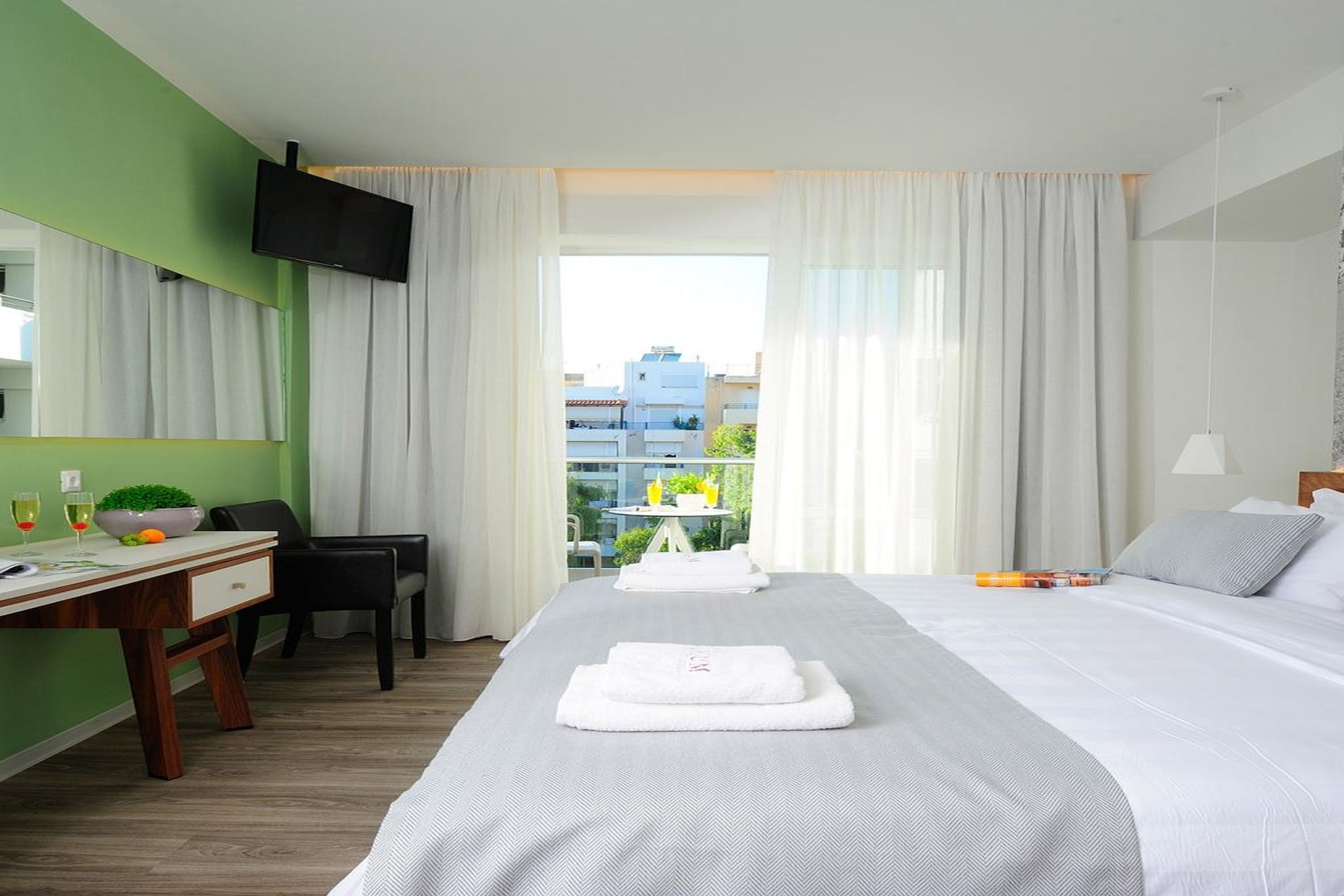 Habitación de hotel con cama, escritorio, silla, televisor y balcón con vista a edificios.