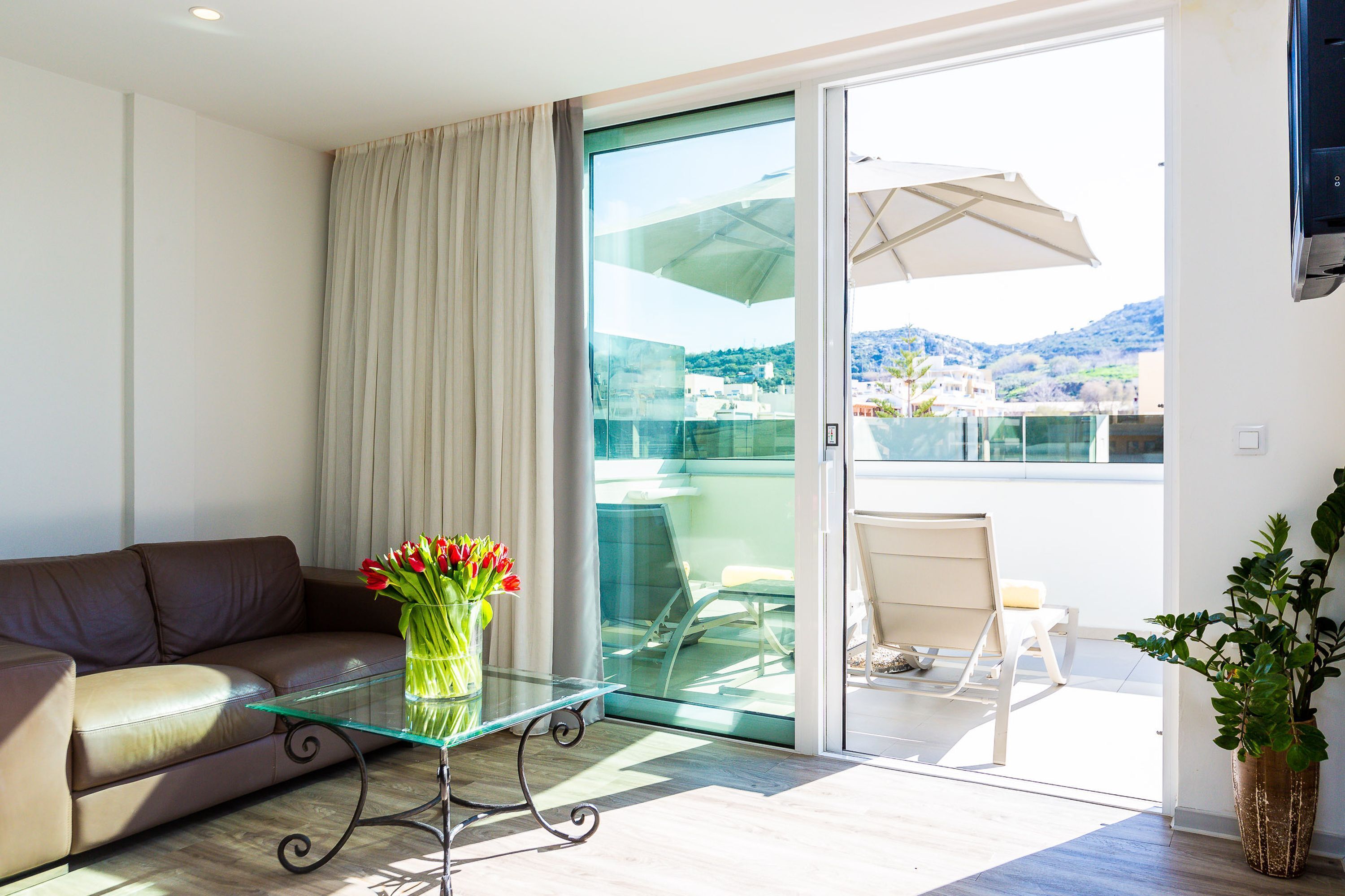 A modern living room with a glass coffee table, a brown leather sofa, a vase with red flowers, and a sliding glass door leading to a balcony with a white umbrella and a chair.