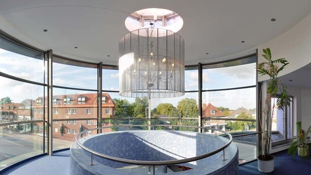 A modern interior with a circular seating area, large windows offering views of buildings and trees, and a hanging light fixture. - Hotel-Restaurant Thomsen (Delmenhorst)