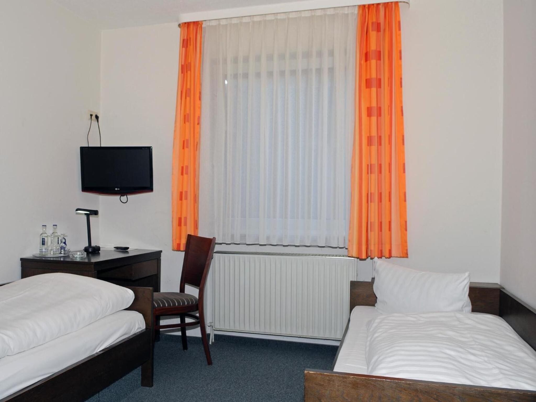 A hotel room with two beds, a desk with a computer, a chair, a TV, and a window with orange curtains.