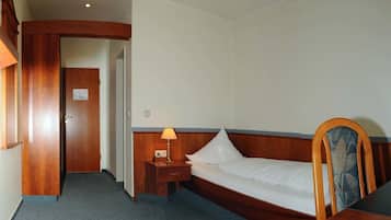 Habitación de hotel pequeña con una cama individual, una mesita de noche con lámpara, cabecera de madera y una puerta que da a otra habitación.