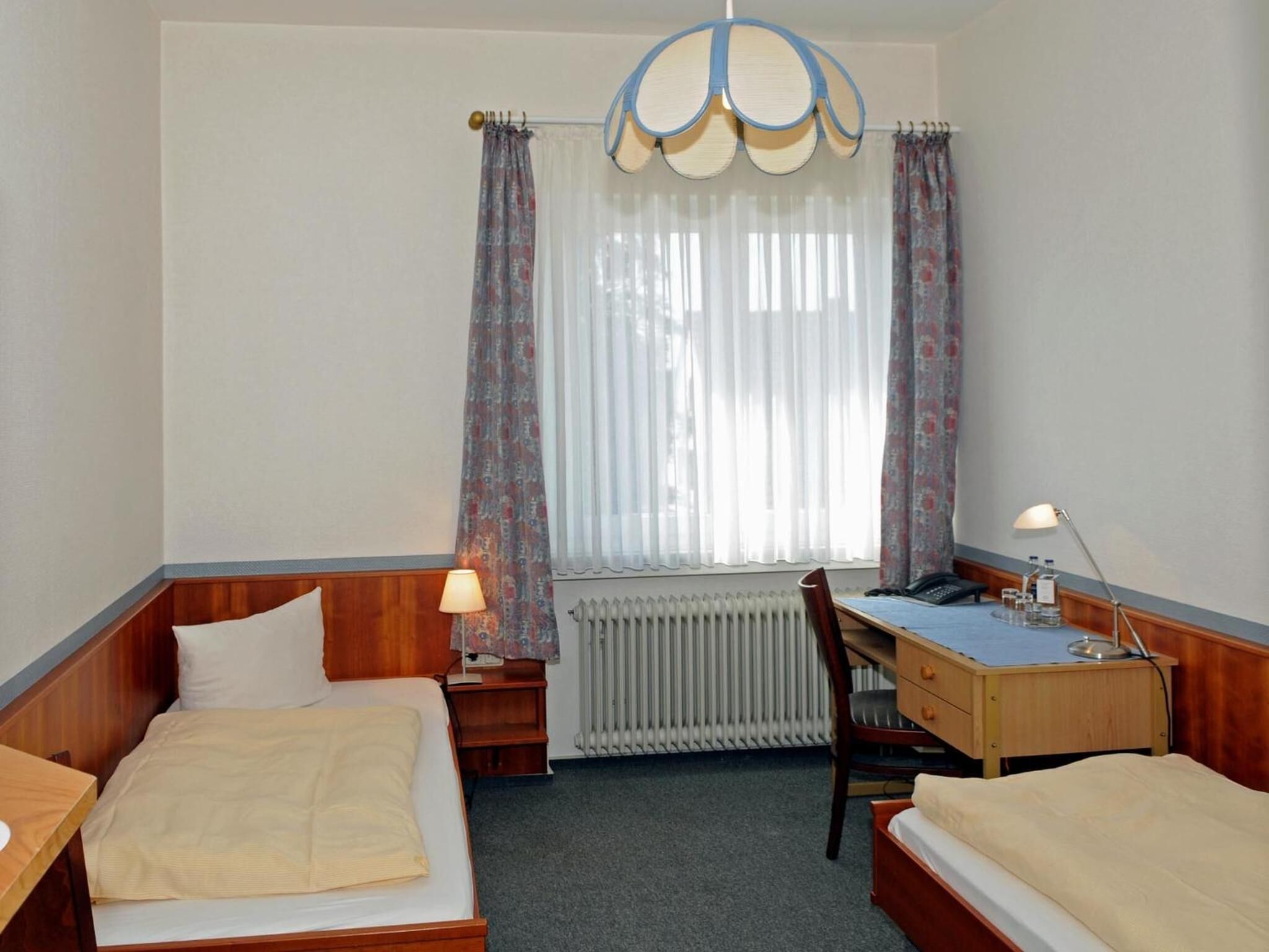A hotel room with two beds, a desk, a chair, a radiator, and a window with curtains.