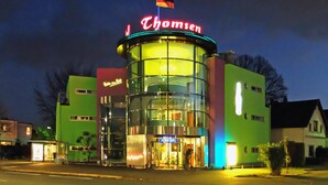 A modern hotel with a glass facade, a neon sign, and a flag on top. - Hotel-Restaurant Thomsen (Delmenhorst)