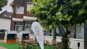 A hotel exterior with a green outdoor seating area, a large potted plant, and a flag with a logo. - Hotel-Restaurant Thomsen (Delmenhorst)