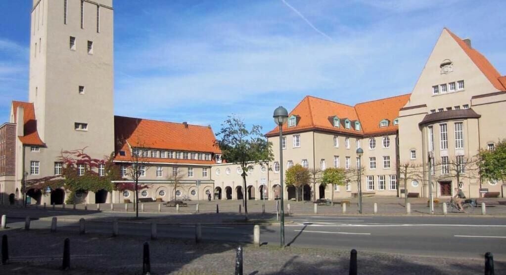 Un bâtiment historique doté d’une tour imposante, d’une cour avec des bancs et d’une rue qui s’étend devant lui.