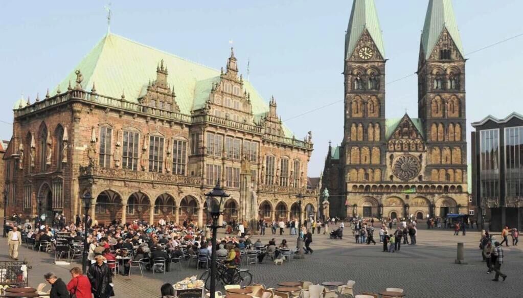 A historic building with a prominent clock tower and a large open plaza with outdoor seating.