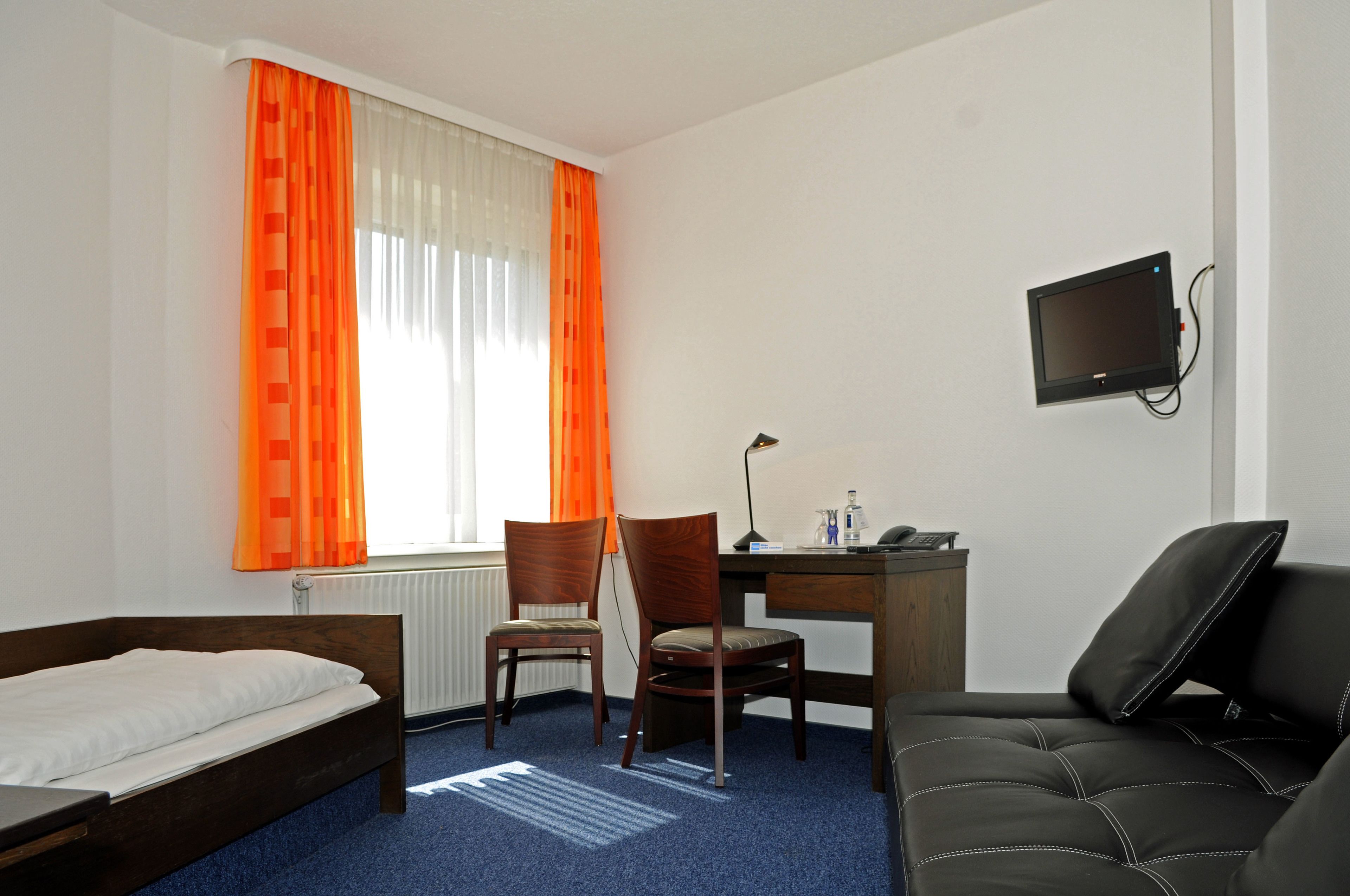 Habitación de hotel con una cama, dos sillas de madera, un escritorio con una computadora, un televisor montado en la pared y una ventana con cortinas naranjas.