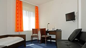 Habitación de hotel con una cama, dos sillas de madera, un escritorio con una computadora, un televisor montado en la pared y una ventana con cortinas naranjas.