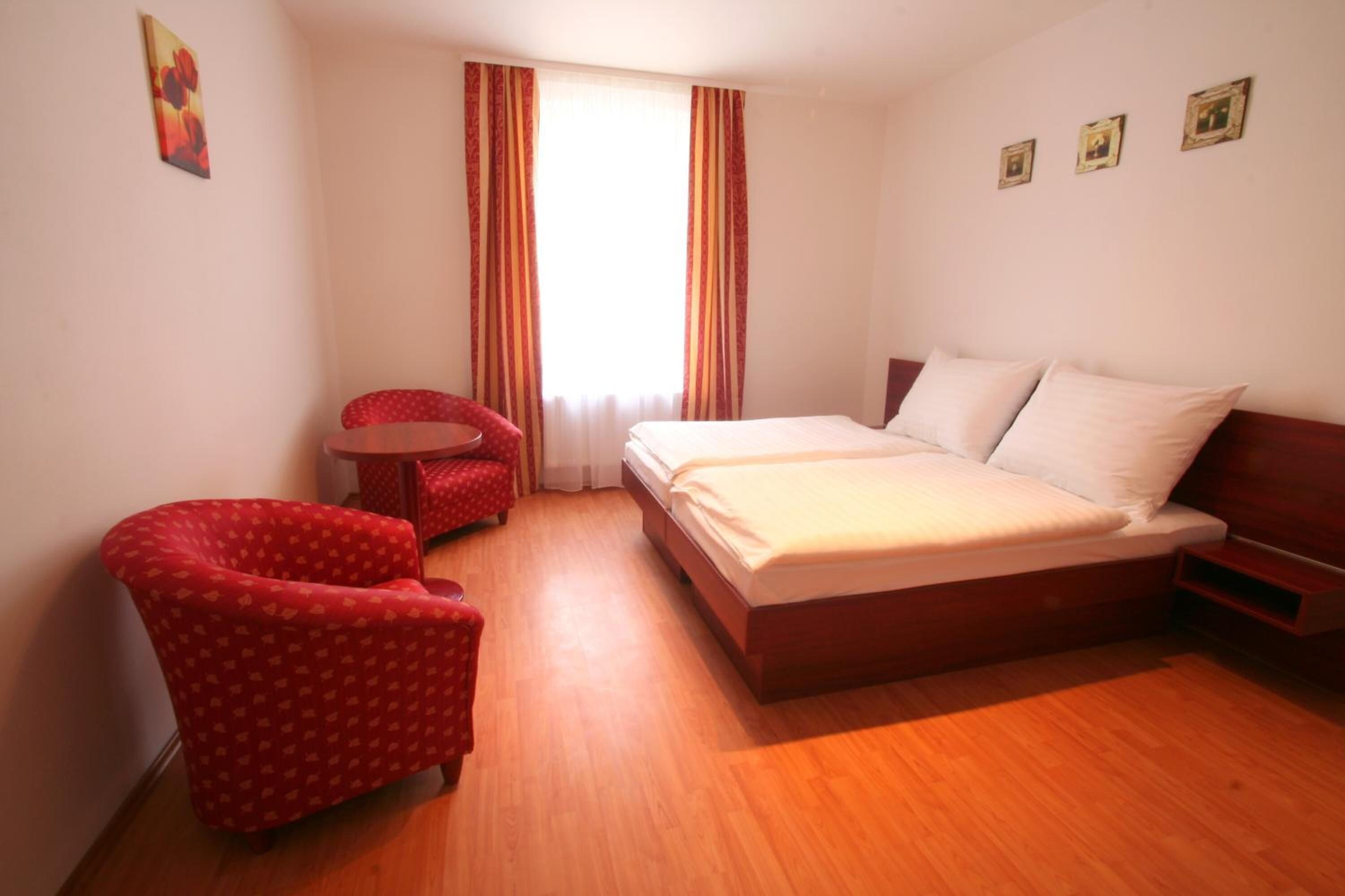 Una camera d'albergo con pavimento in legno, un letto con biancheria bianca, una poltrona rossa e un tavolino.