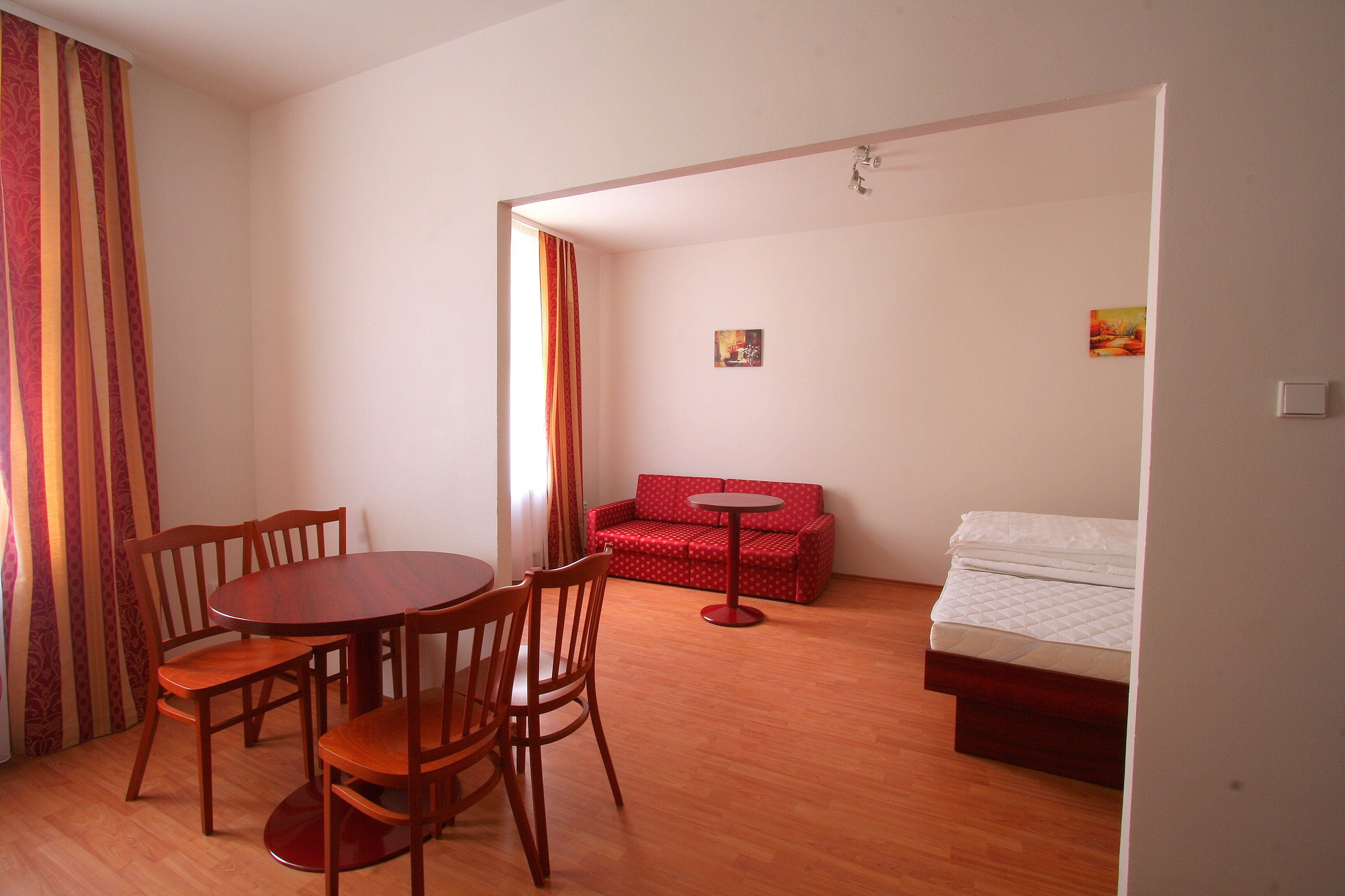 A room with a wooden dining table and chairs, a red sofa, a small round table, and a bed with a white mattress.