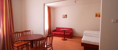A room with a wooden dining table and chairs, a red sofa, a small round table, and a bed with a white mattress.