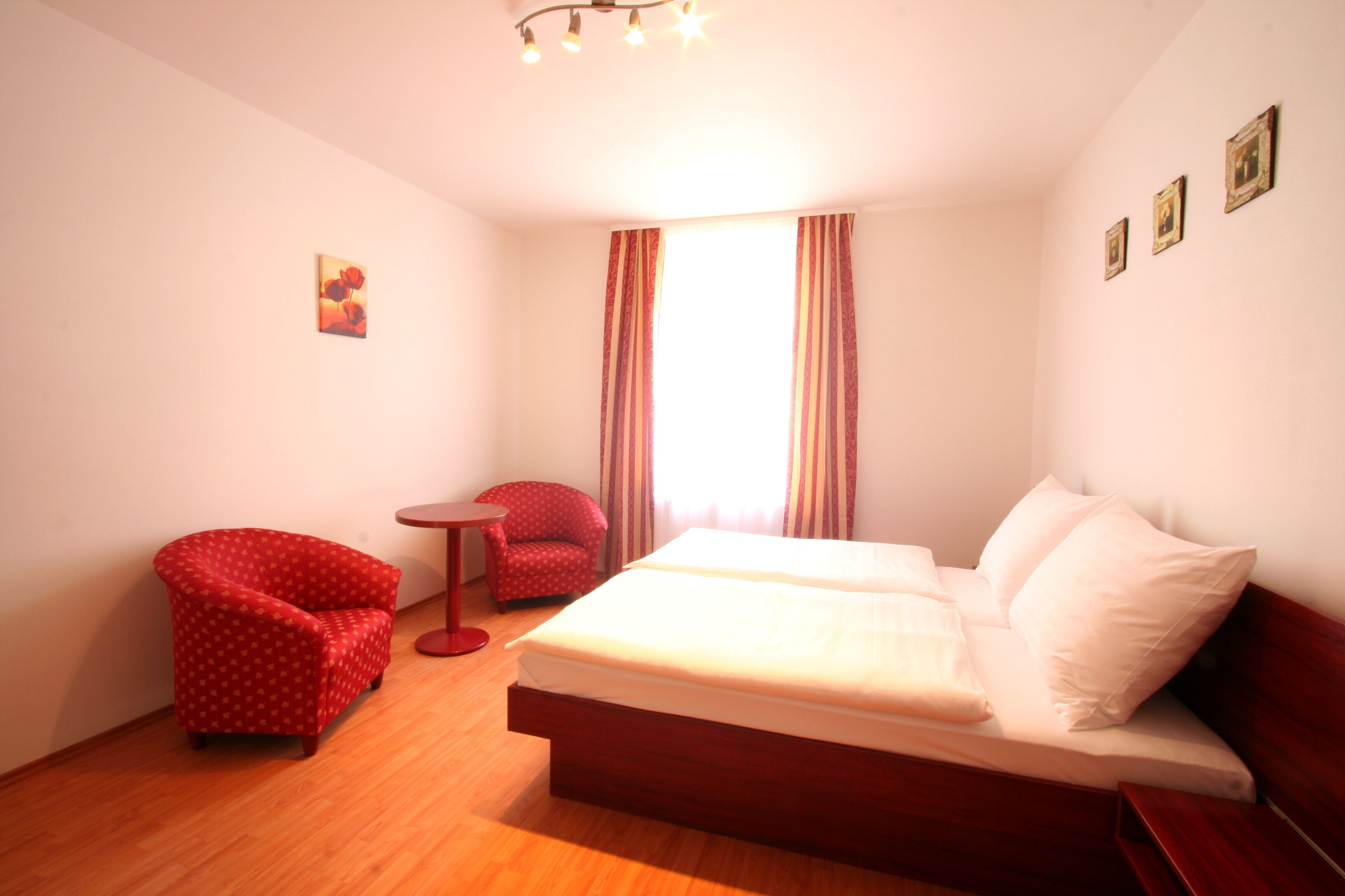 A hotel room with a wooden floor, a single bed with white bedding, a red armchair, a small round table, and a window with curtains.