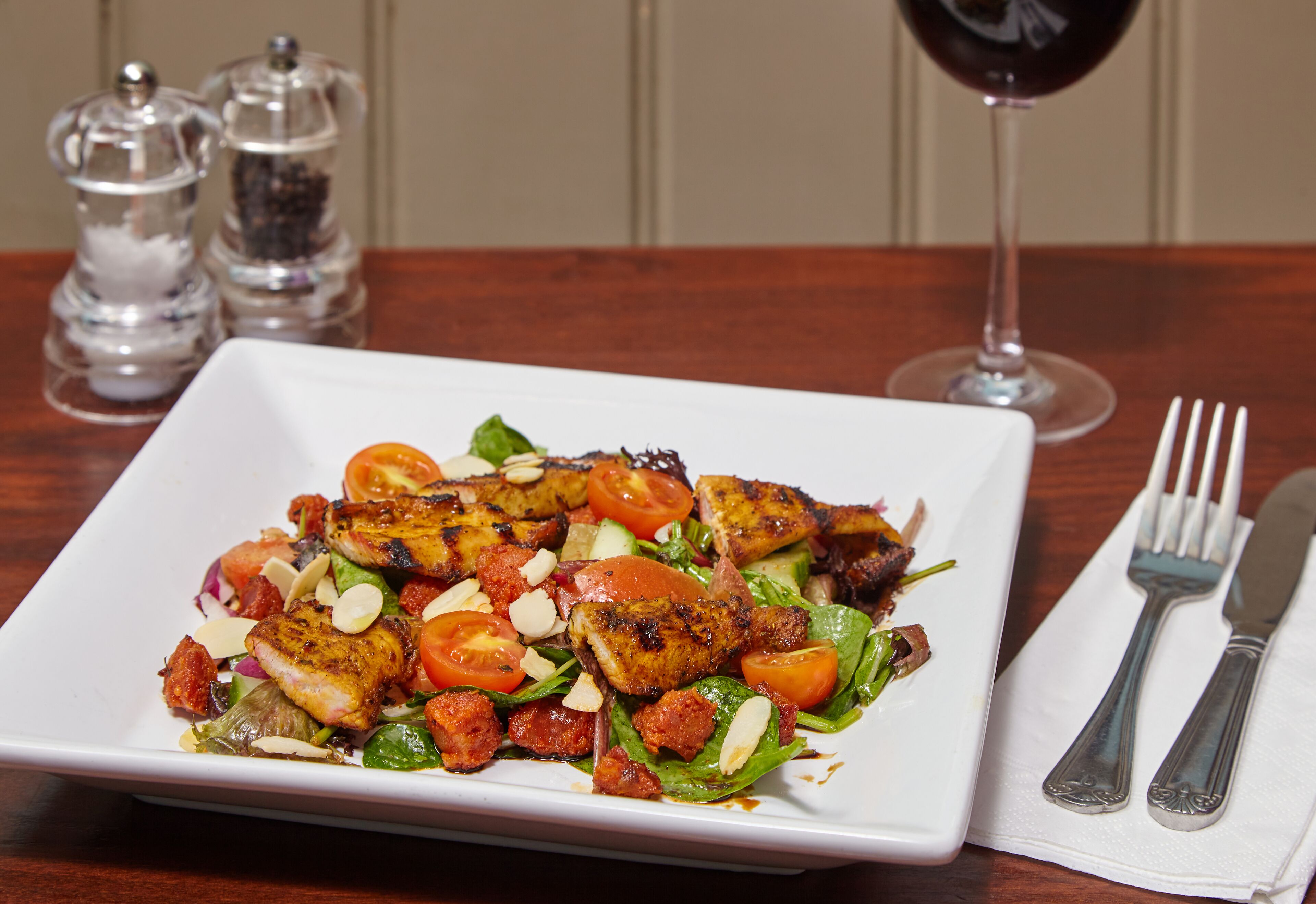 A square white plate with grilled chicken, tomatoes, and greens, accompanied by a glass of red wine and cutlery on a wooden table.
