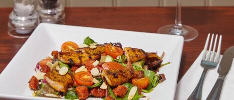 A square white plate with grilled chicken, tomatoes, and greens, accompanied by a glass of red wine and cutlery on a wooden table.
