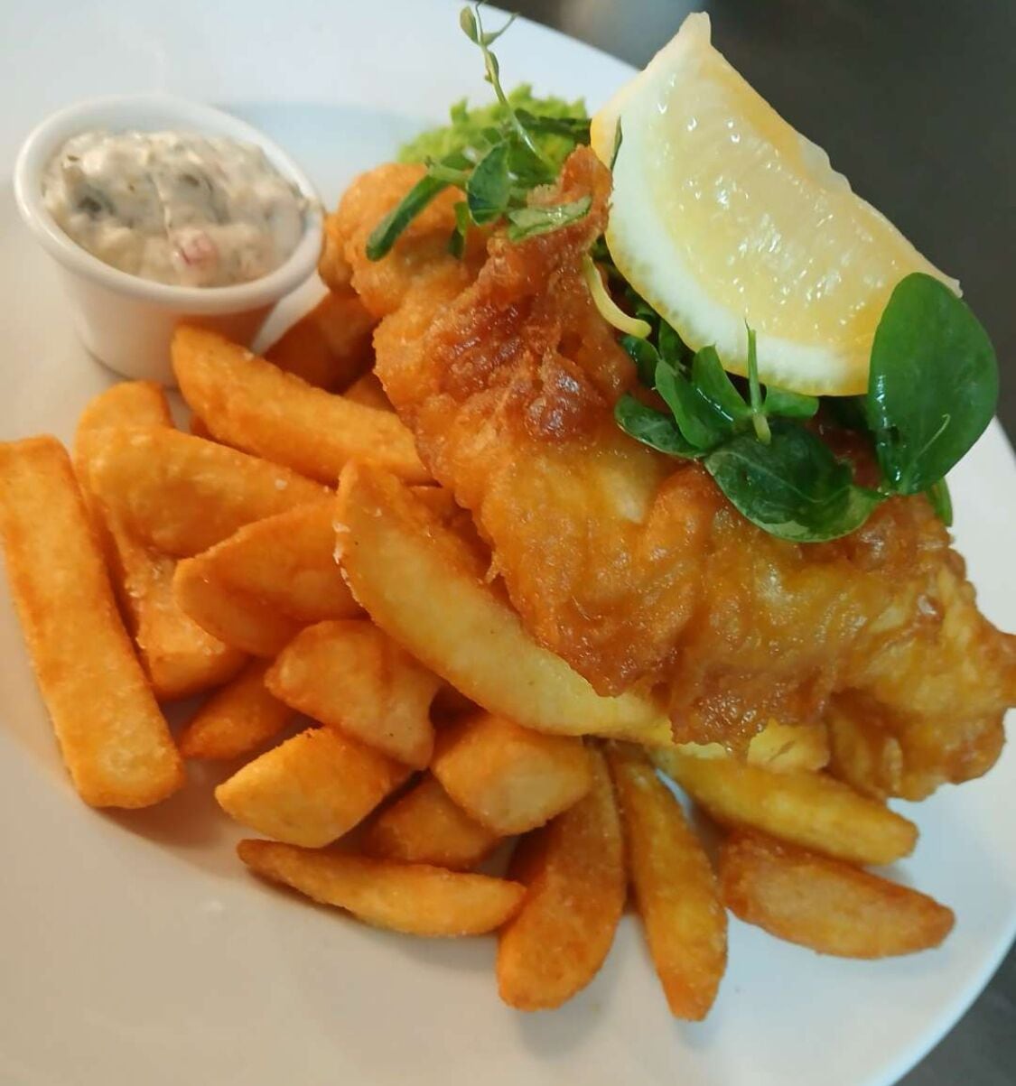 Un plato de pescado y papas fritas acompañado de salsa tártara y un gajo de limón.