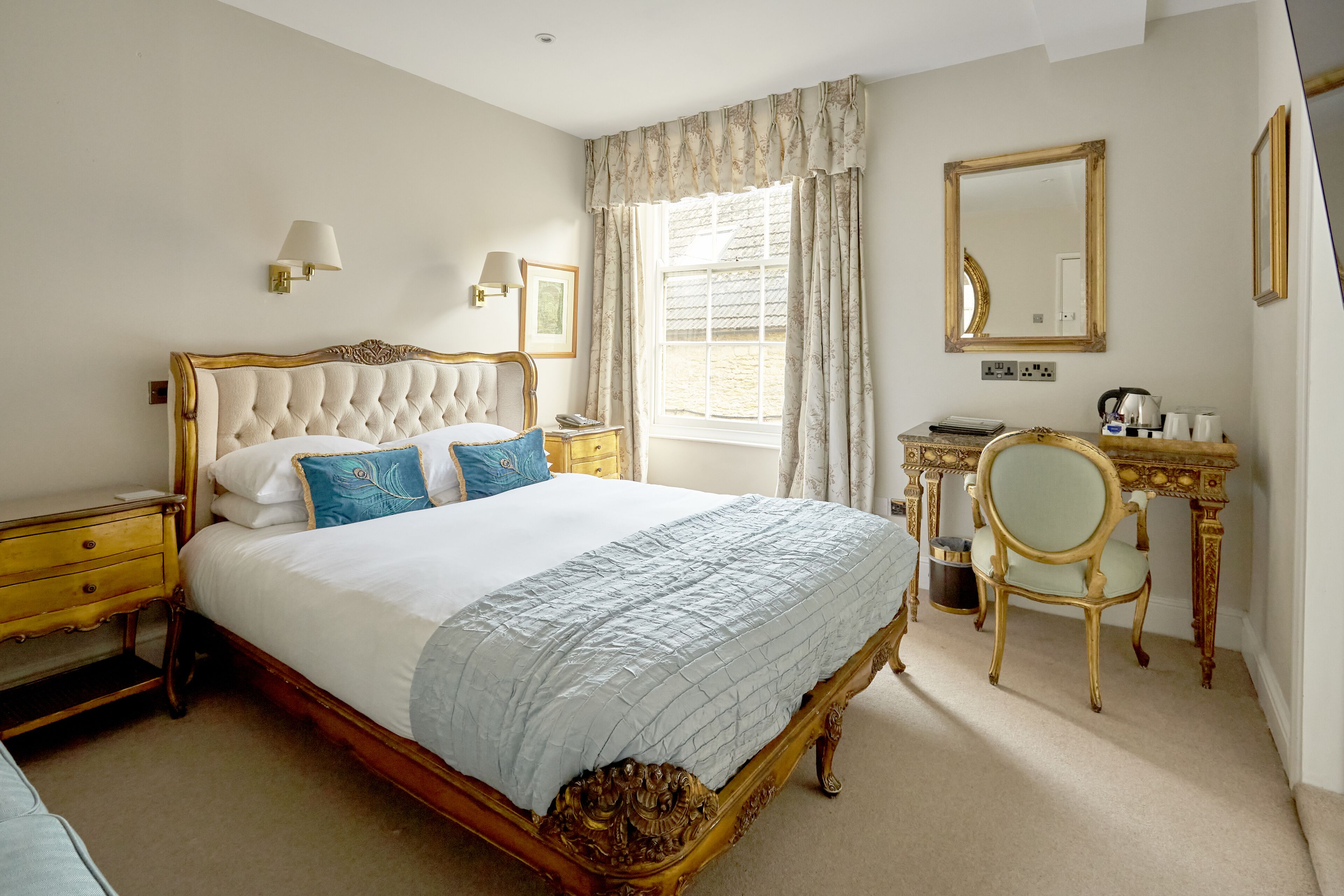 A classic bedroom with a large bed, a wooden nightstand, a desk with a chair, a mirror, and a window with curtains.