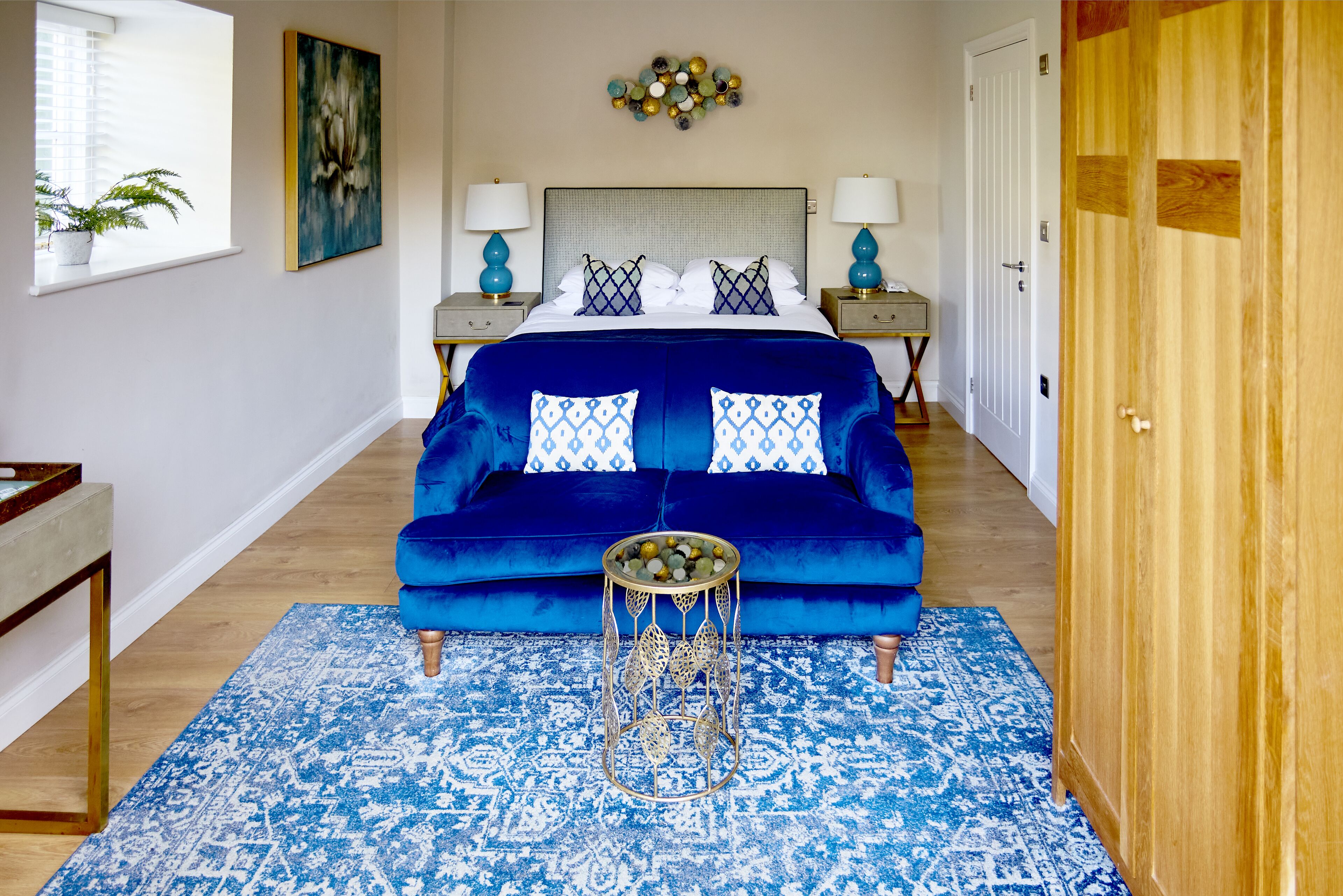 Un dormitorio con un sofá de terciopelo azul, un tapete estampado, una mesita de noche con lámpara y un adorno de pared.