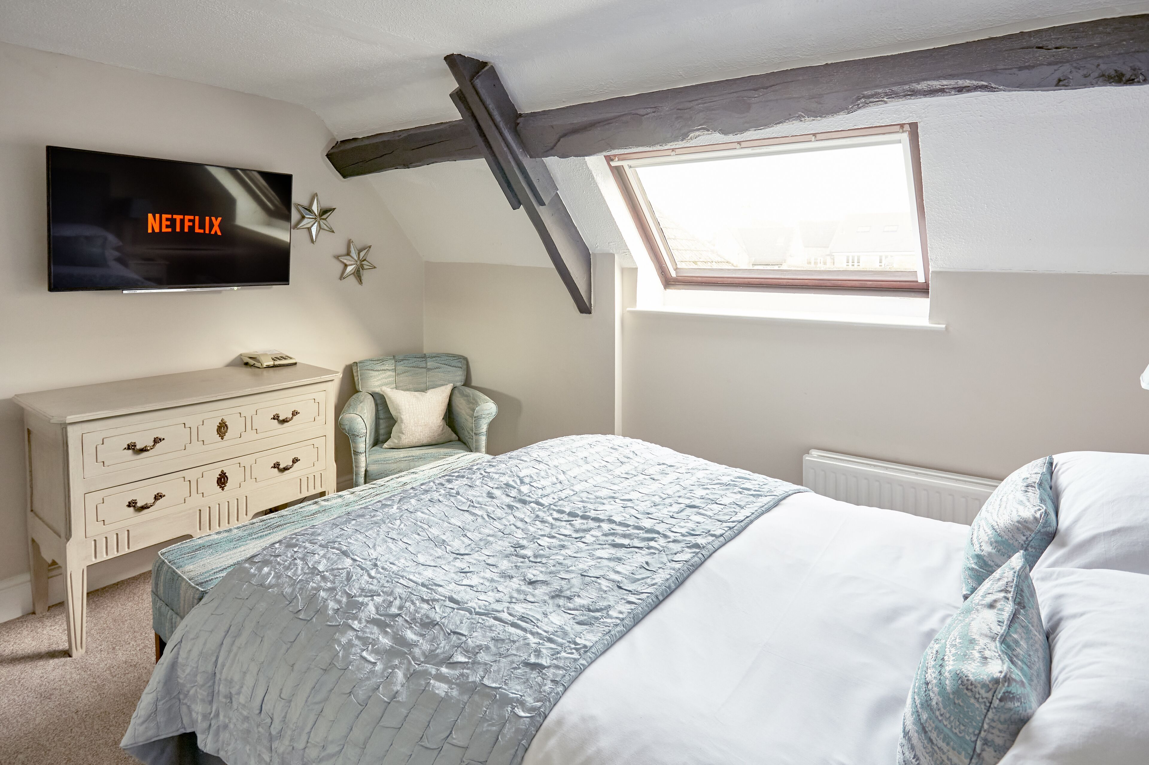 Un dormitorio con cama, cómoda, silla, televisor montado en la pared y tragaluz.