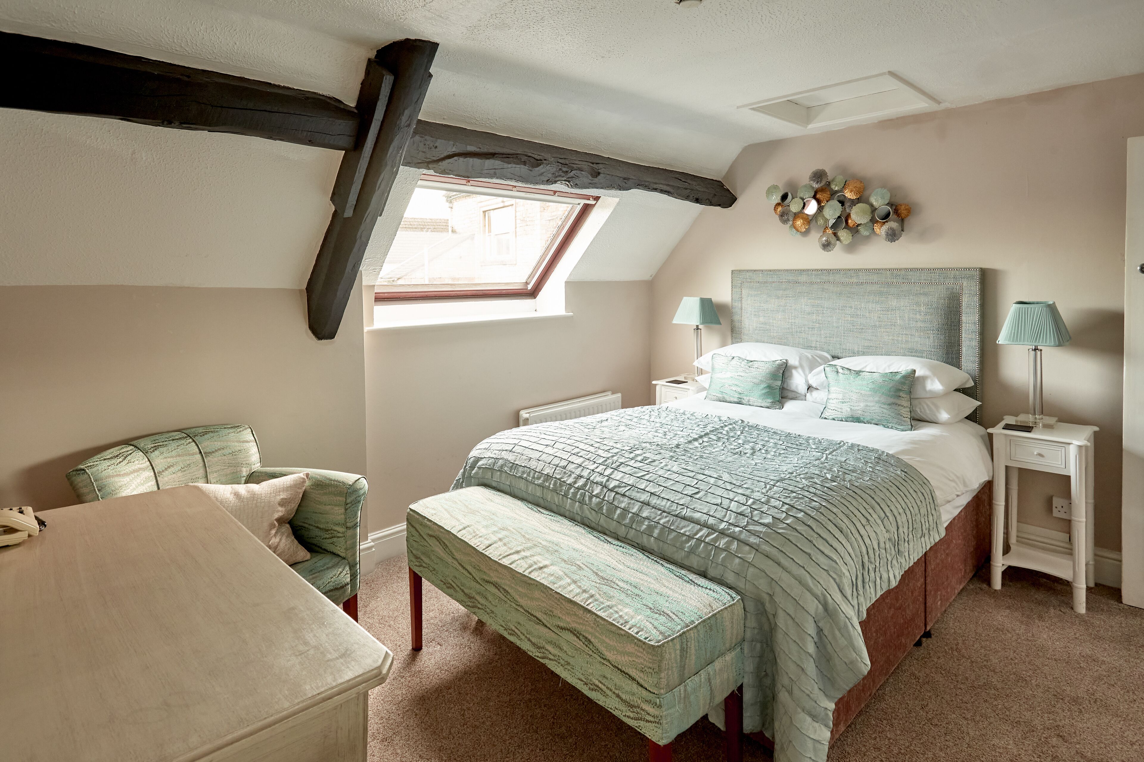 A bedroom with a sloped ceiling, a skylight, a bed with a headboard, bedside tables, a chair, and a bench.