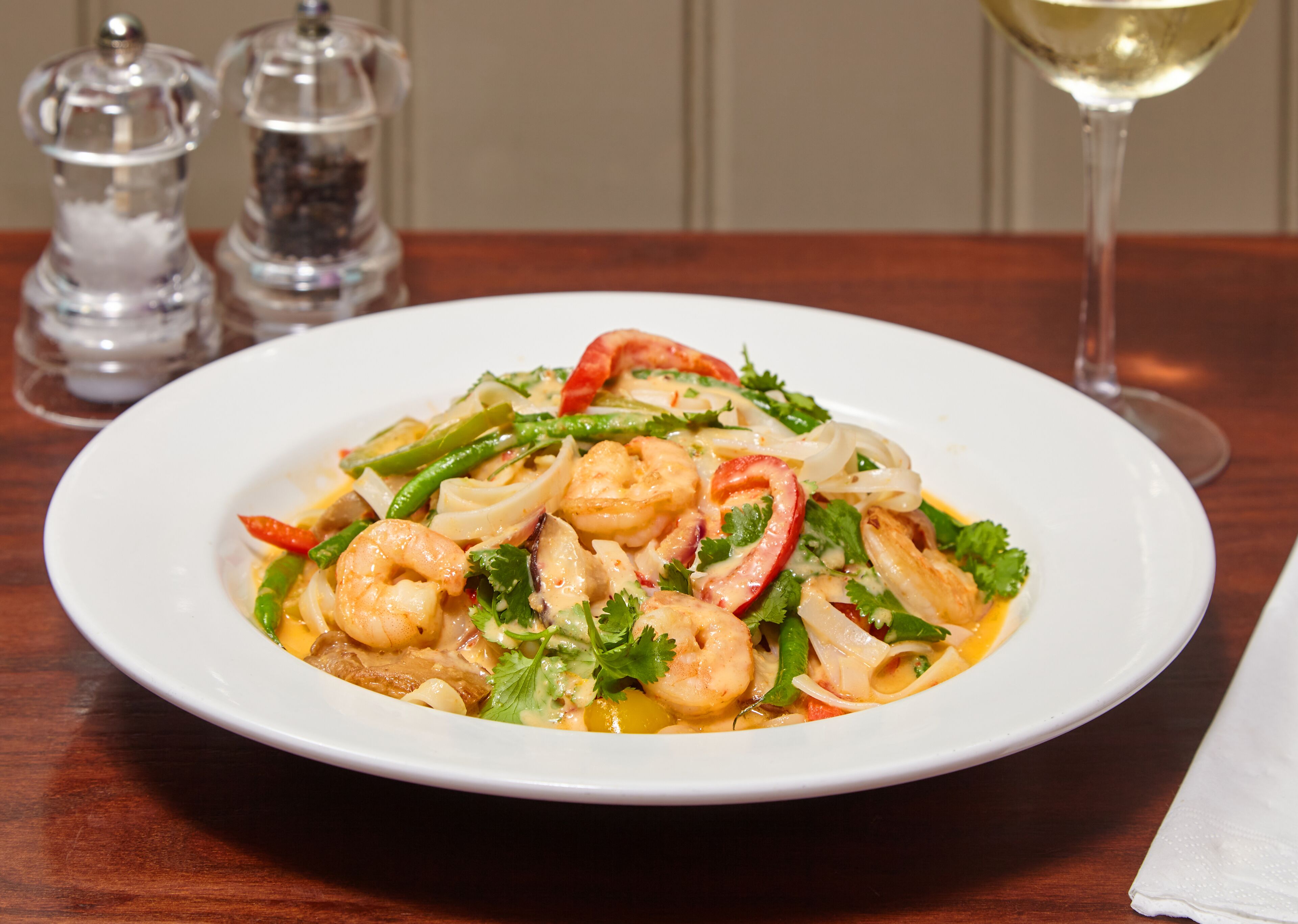 A plate of shrimp and vegetables on a wooden table with salt and pepper shakers and a wine glass in the background.