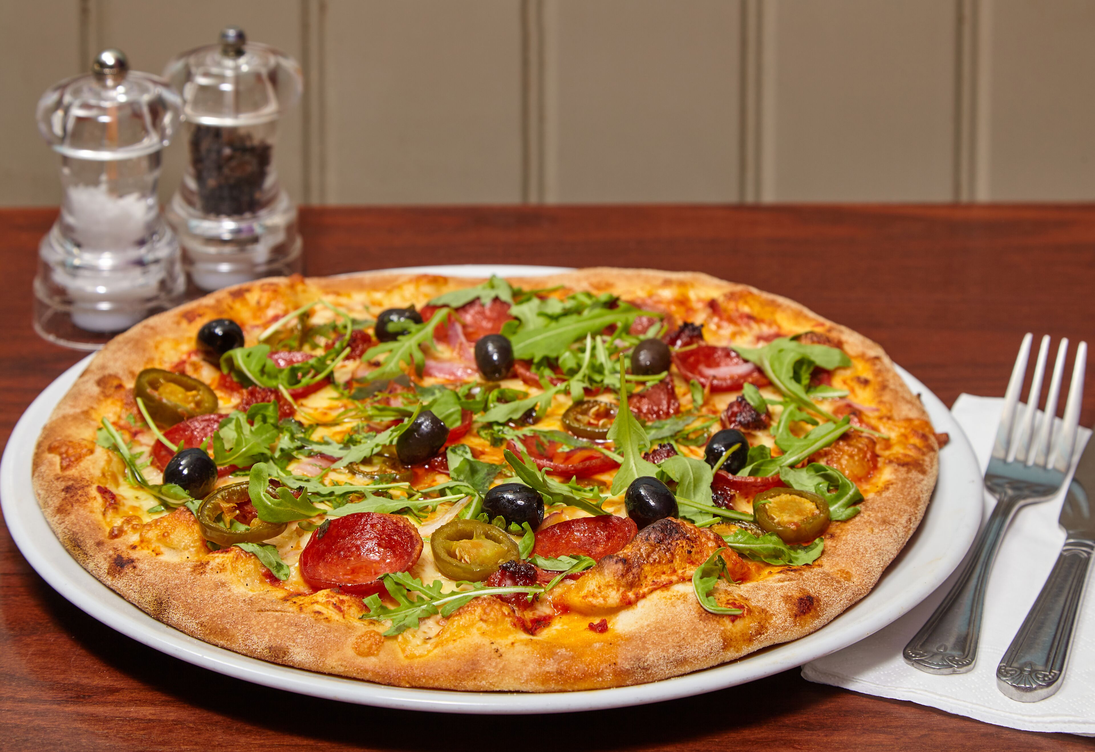 A pizza with various toppings on a white plate, placed on a wooden table with salt and pepper shakers in the background.