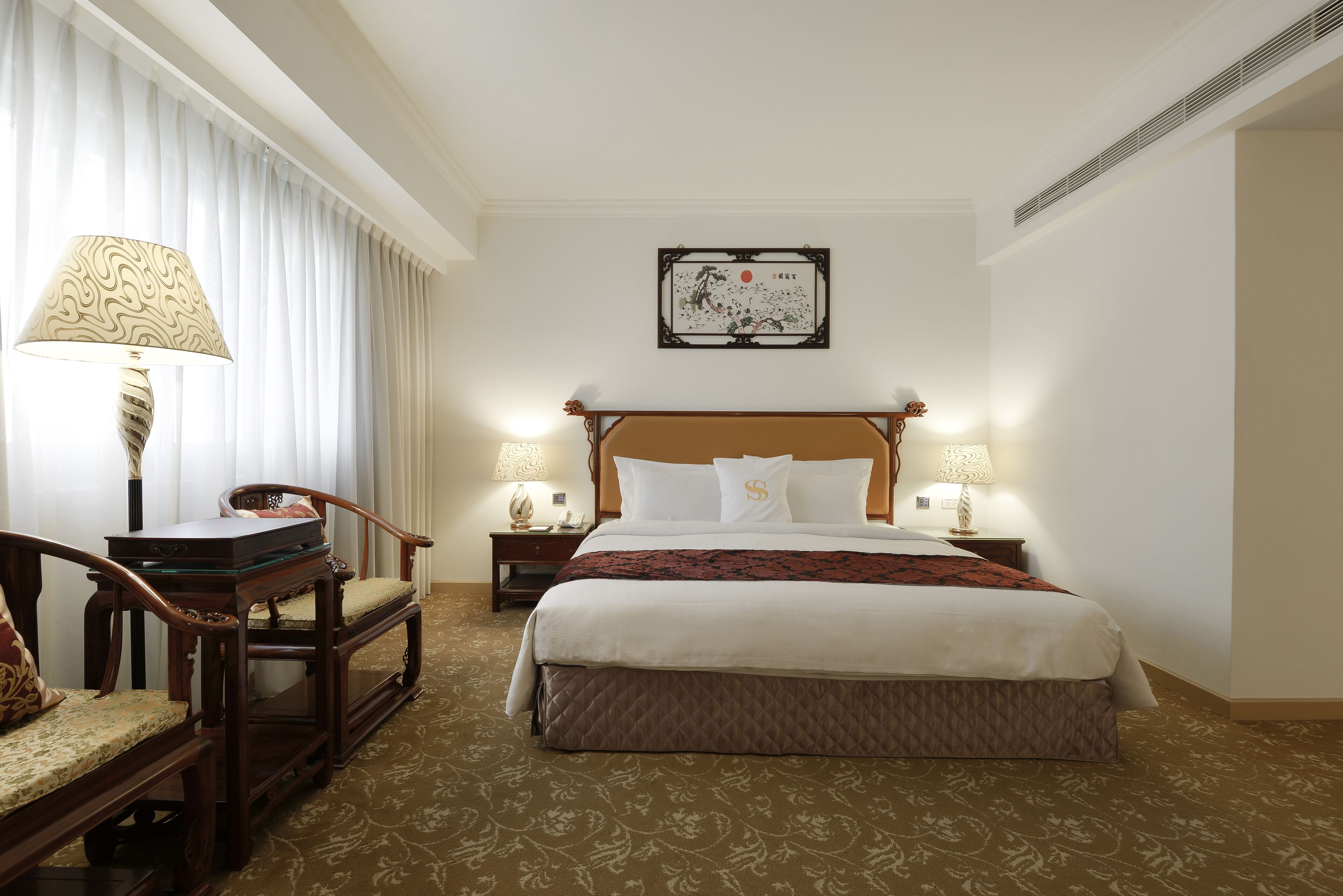 Una habitación de hotel con una cama grande, dos mesitas de noche con lámparas, una silla de madera y un adorno en la pared.