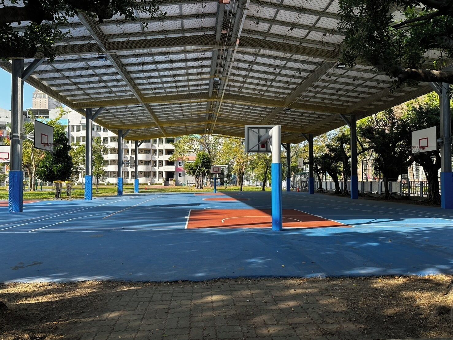 Una cancha de baloncesto al aire libre con zona de estar techada, rodeada de árboles y edificios.