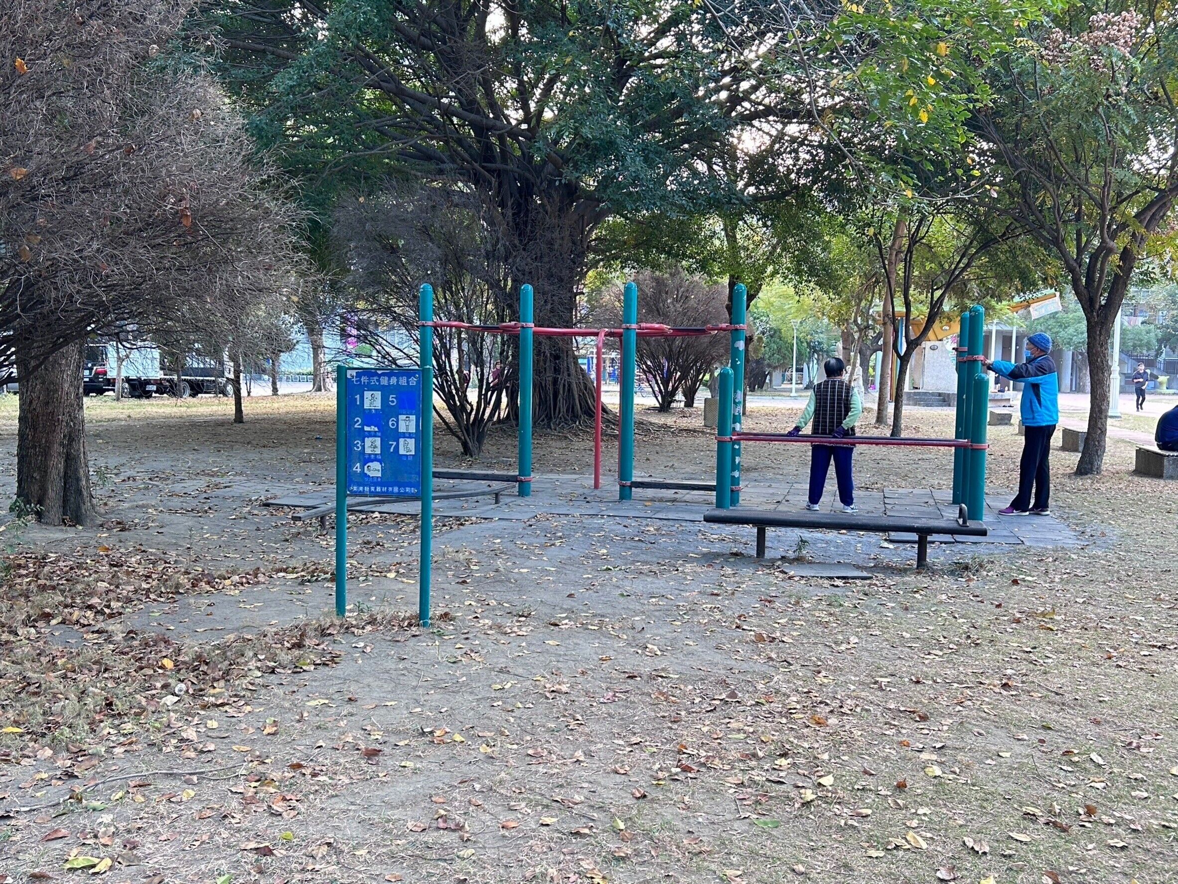 Un parc amb aparells d'exercici, un cartell blau amb símbols i arbres.