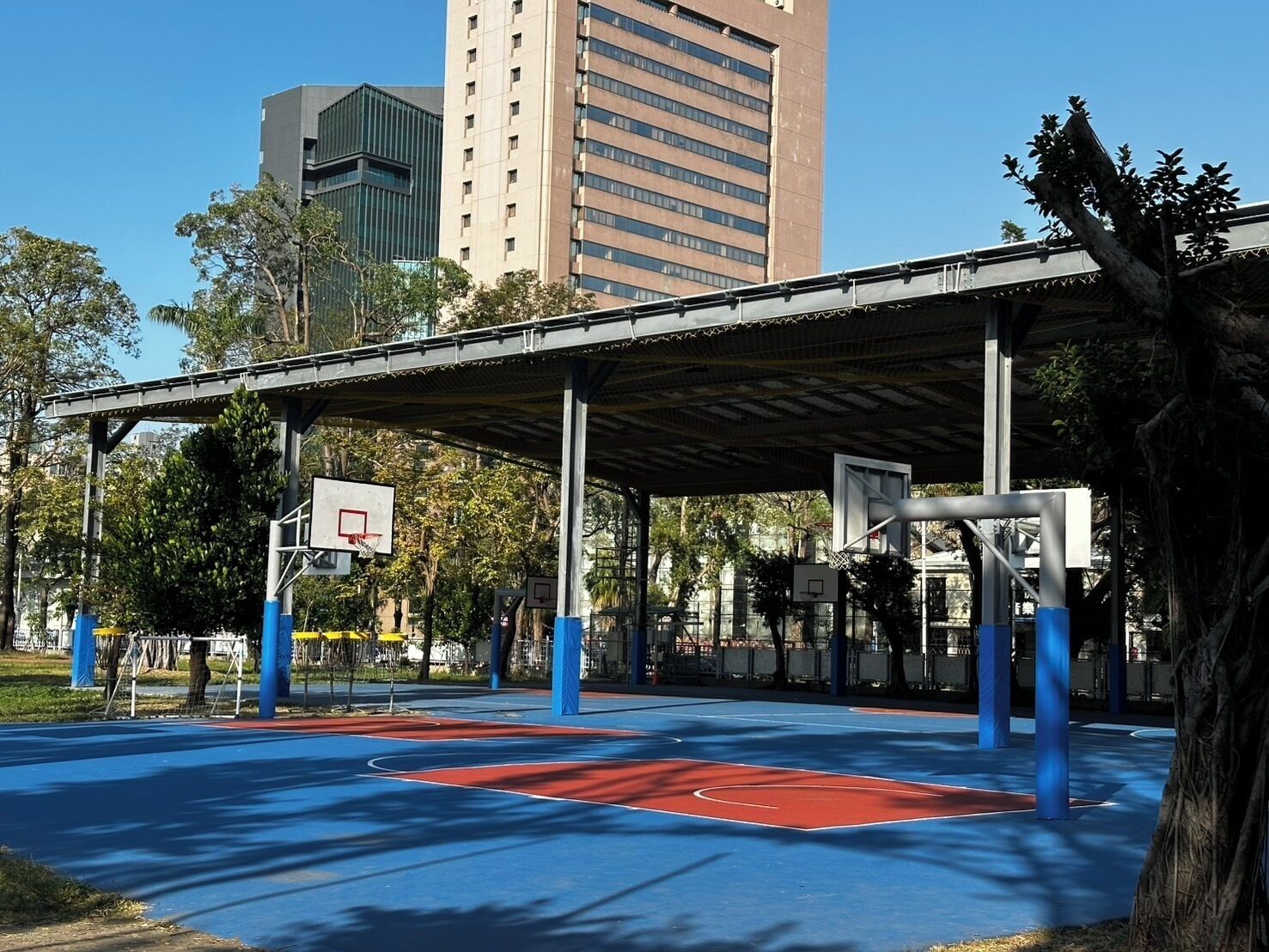 An outdoor basketball court with a covered seating area, surrounded by trees and buildings.