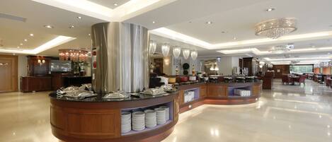A modern hotel lobby with a central buffet area, wooden paneling, and a chandelier.