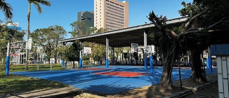 An outdoor basketball court with a covered seating area, surrounded by trees and buildings.