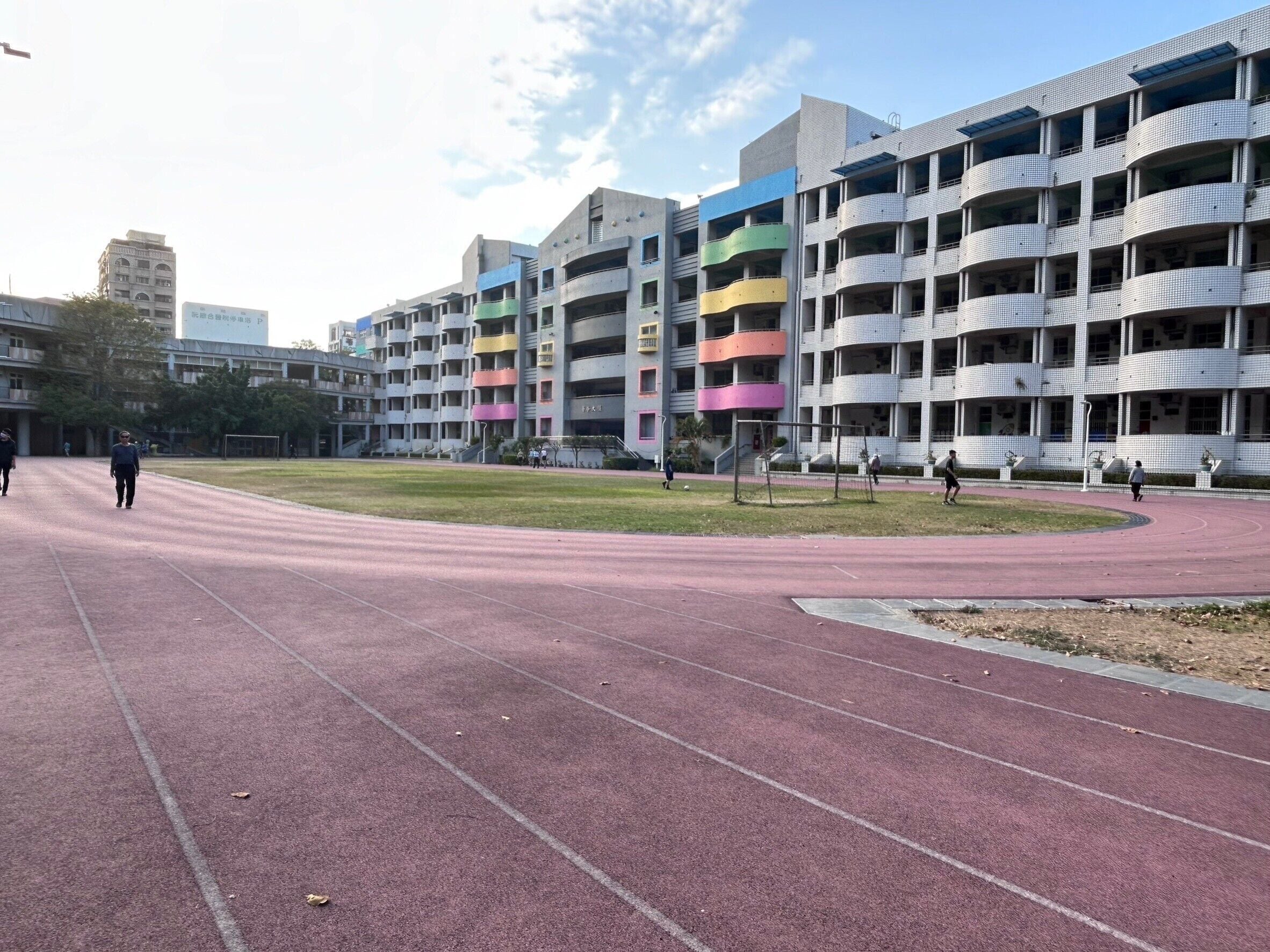 A multi-story building with balconies, a running track, and a grassy area.