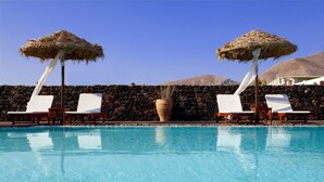 A pool with clear blue water, surrounded by white lounge chairs and thatched umbrellas, with a stone wall and mountains in the background. - Anna Traditional Apartments (Santorini)