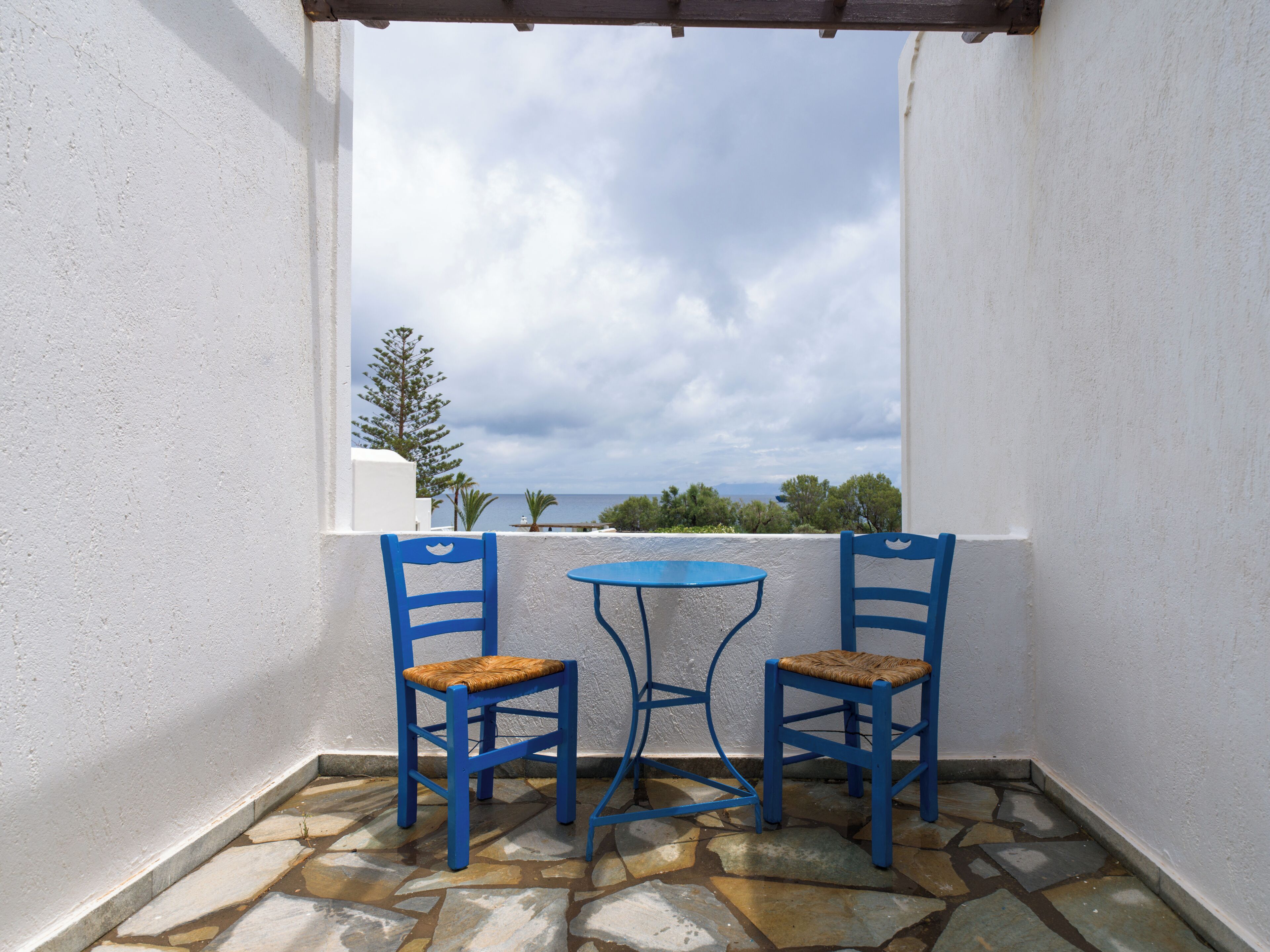 A balcony with two blue chairs and a small table, white walls, and a view of the sea.