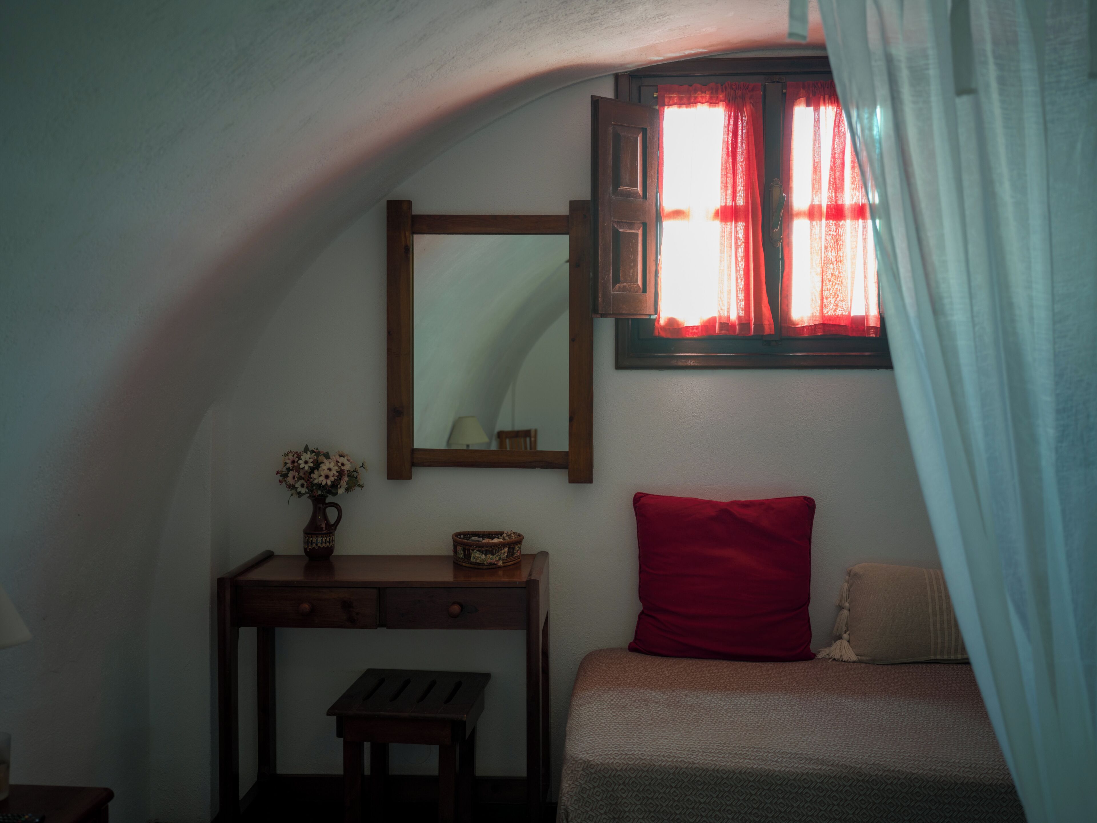 Un dormitorio con techo inclinado, un escritorio de madera con cajones, un espejo, un florero con flores, un cojín rojo y una cama con una manta estampada.