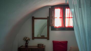 Un dormitorio con techo inclinado, un escritorio de madera con cajones, un espejo, un florero con flores, un cojín rojo y una cama con una manta estampada.