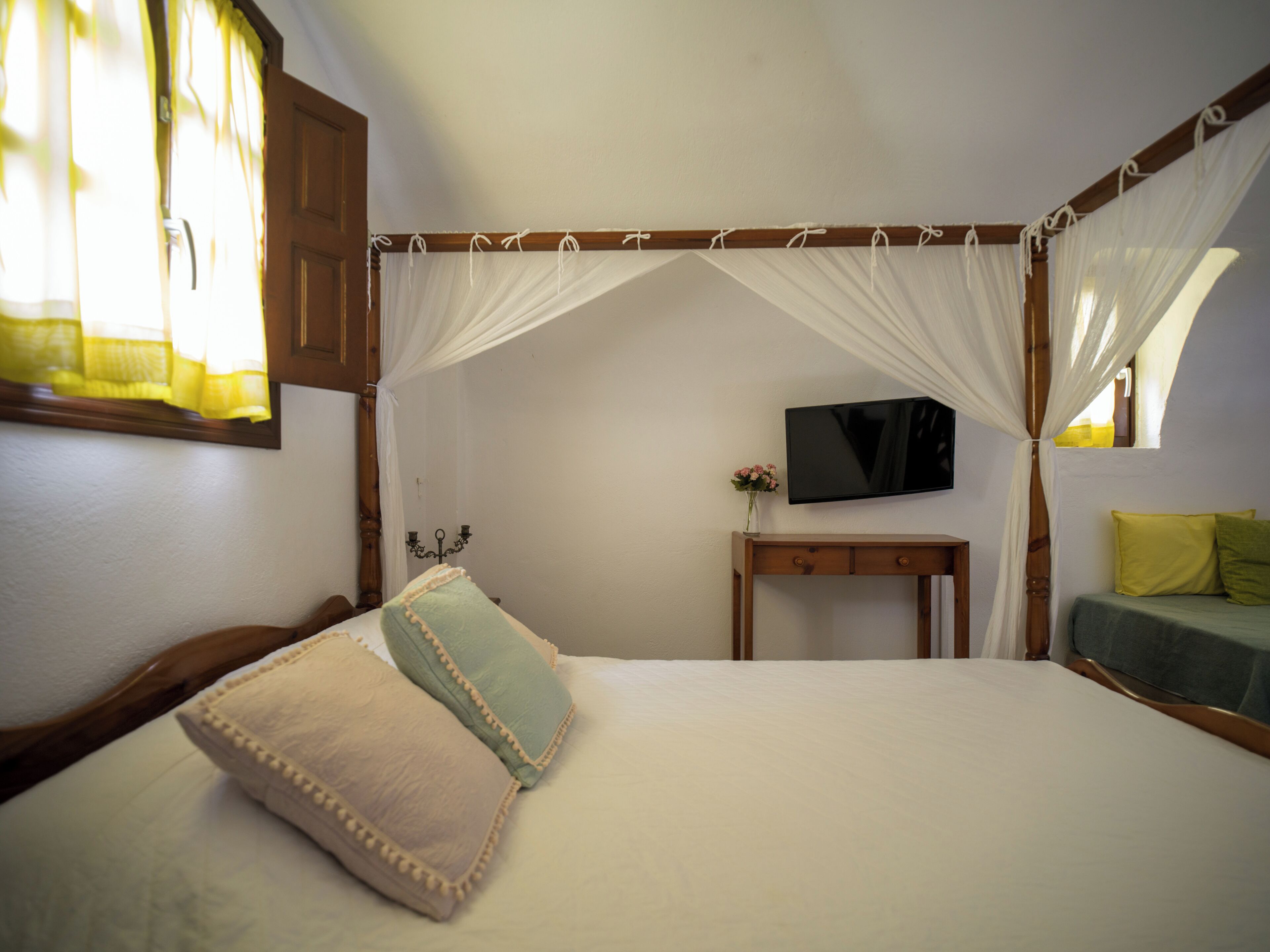 A bedroom with a four-poster bed, a bedside table with a flat-screen TV, and a window with curtains.