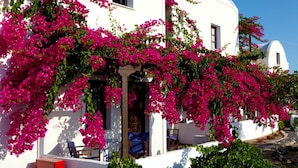A white building with pink bougainvillea vines, a covered outdoor seating area, and a clear blue sky. - Anna Traditional Apartments (Santorini)